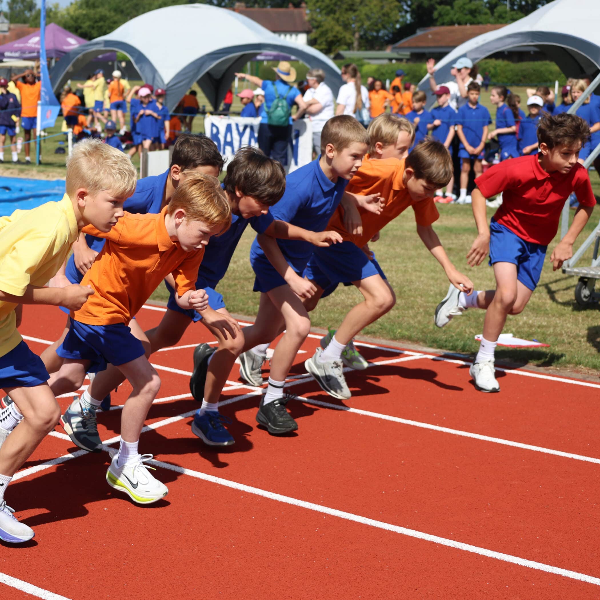 A group of boys in colourful shirts and shorts start a race on a red running track during a school sports day, with spectators and tents visible in the background.
