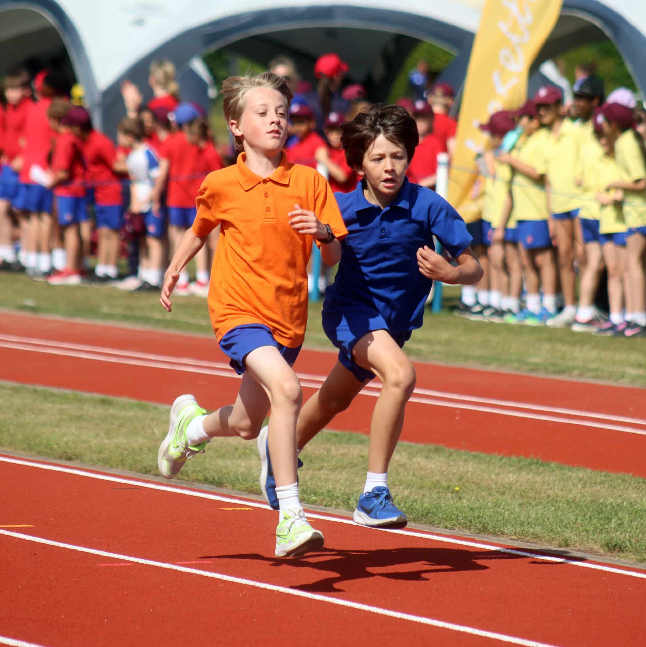 Two boys in sports kits, one in orange and one in blue, race side by side on a running track, with groups of children in colourful kits cheering in the background.