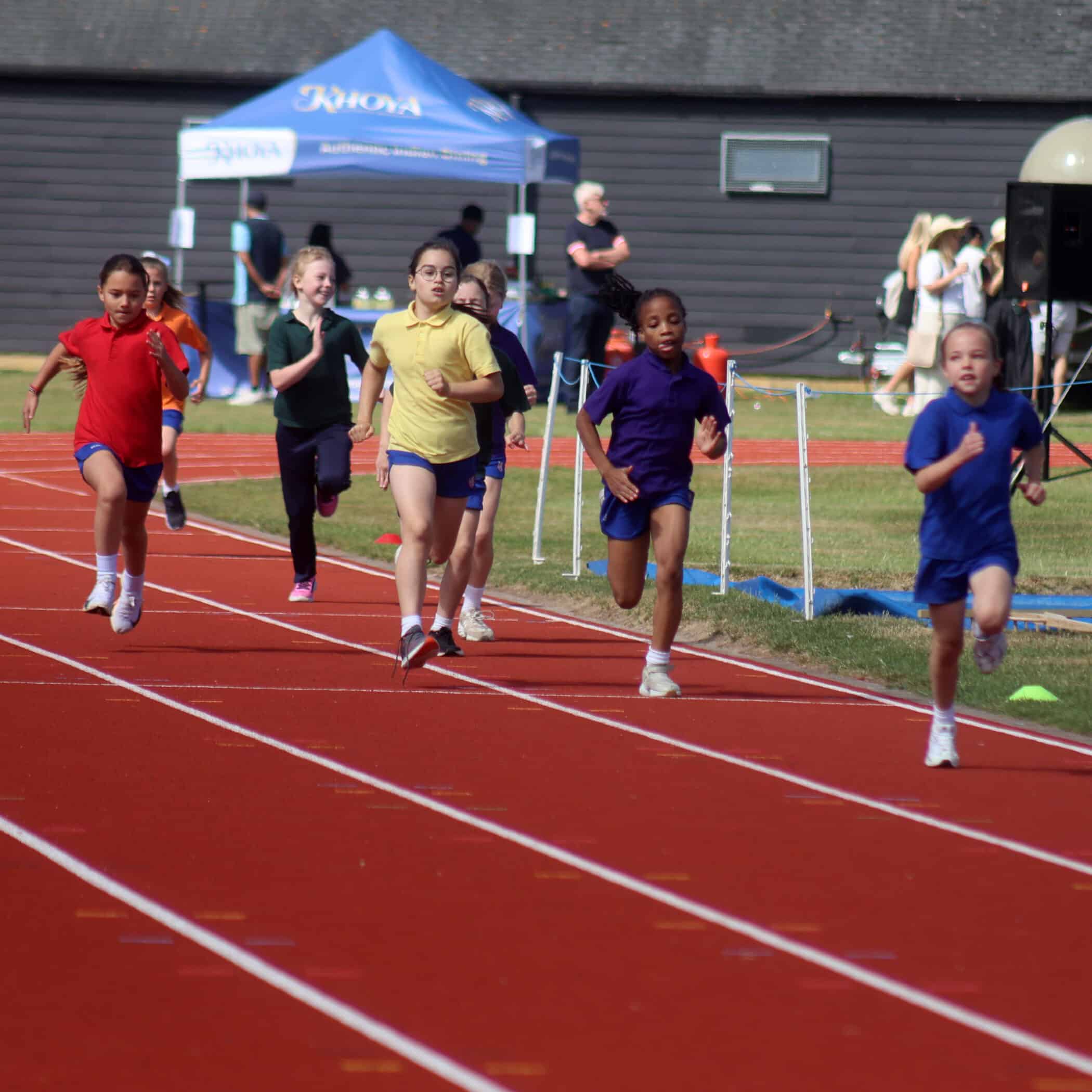 Six young girls run on a red outdoor track during a race, each wearing different coloured shirts. Spectators and a blue marquee are visible in the background on a sunny day.