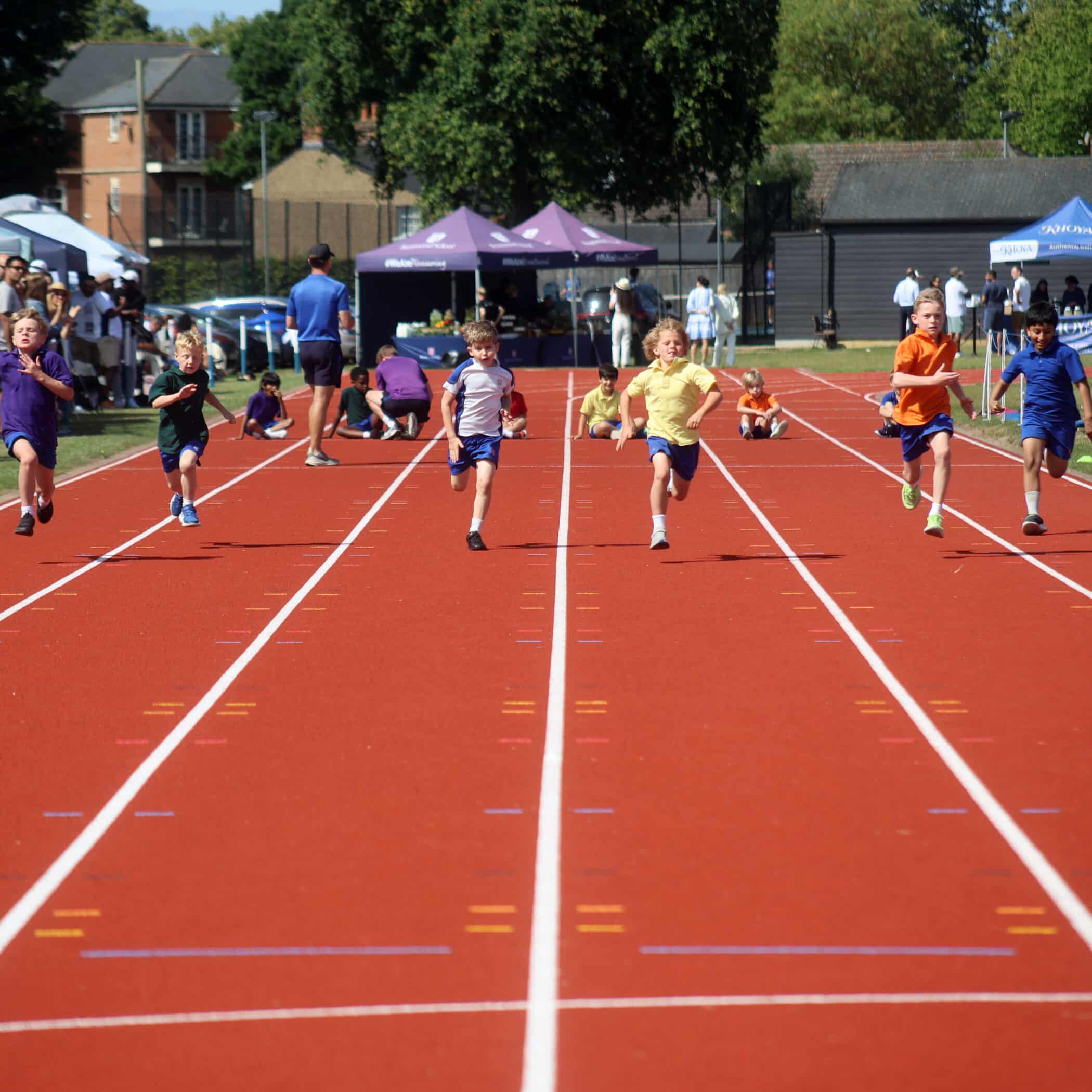 Six young children are sprinting on a red running track during a race on a sunny day, with people and tents in the background near trees and buildings.
