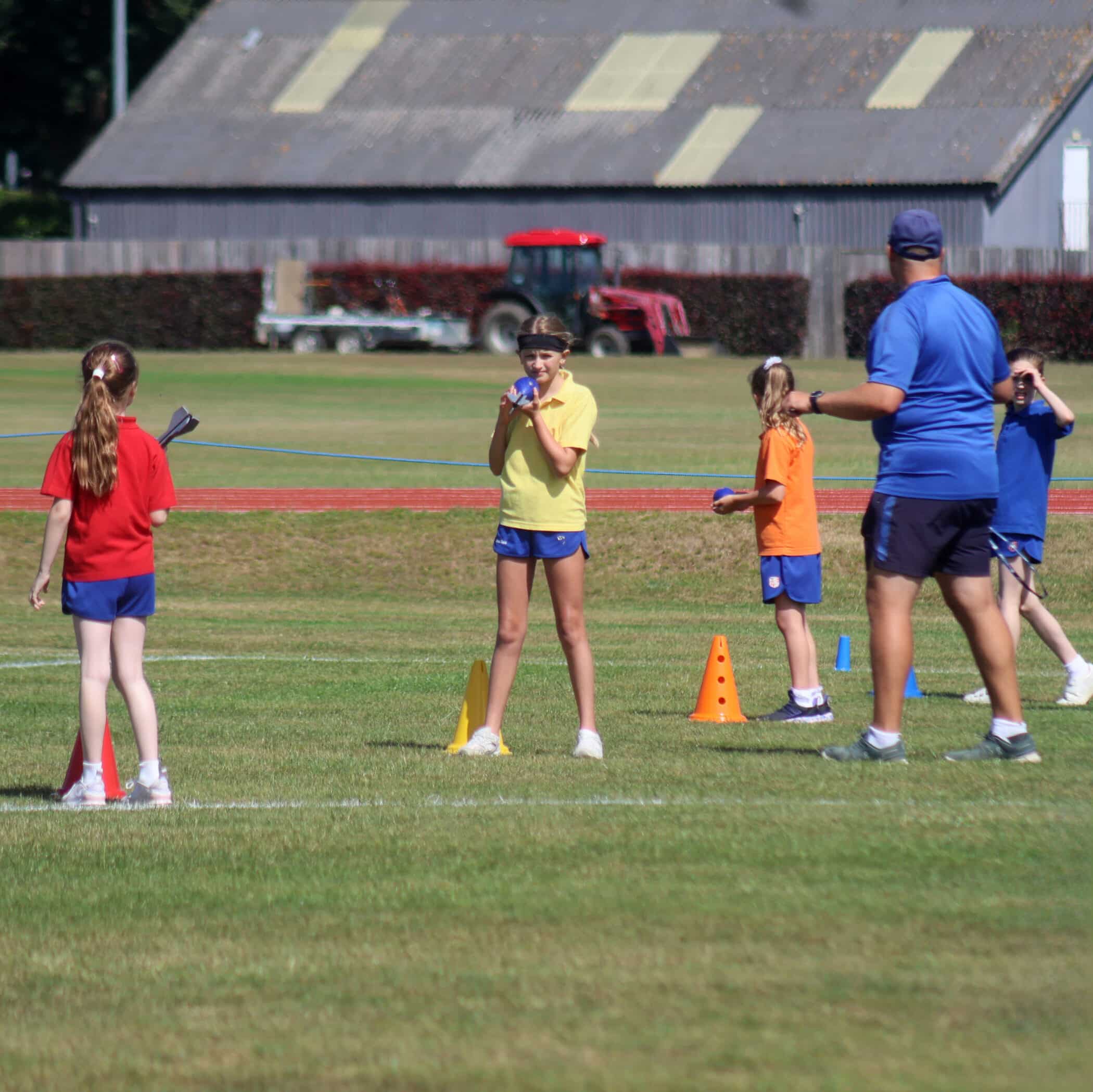 Children in colourful shirts stand by cones on a grassy field, some holding batons. An adult in blue supervises. A barn and red tractor are visible in the background on a sunny day.
