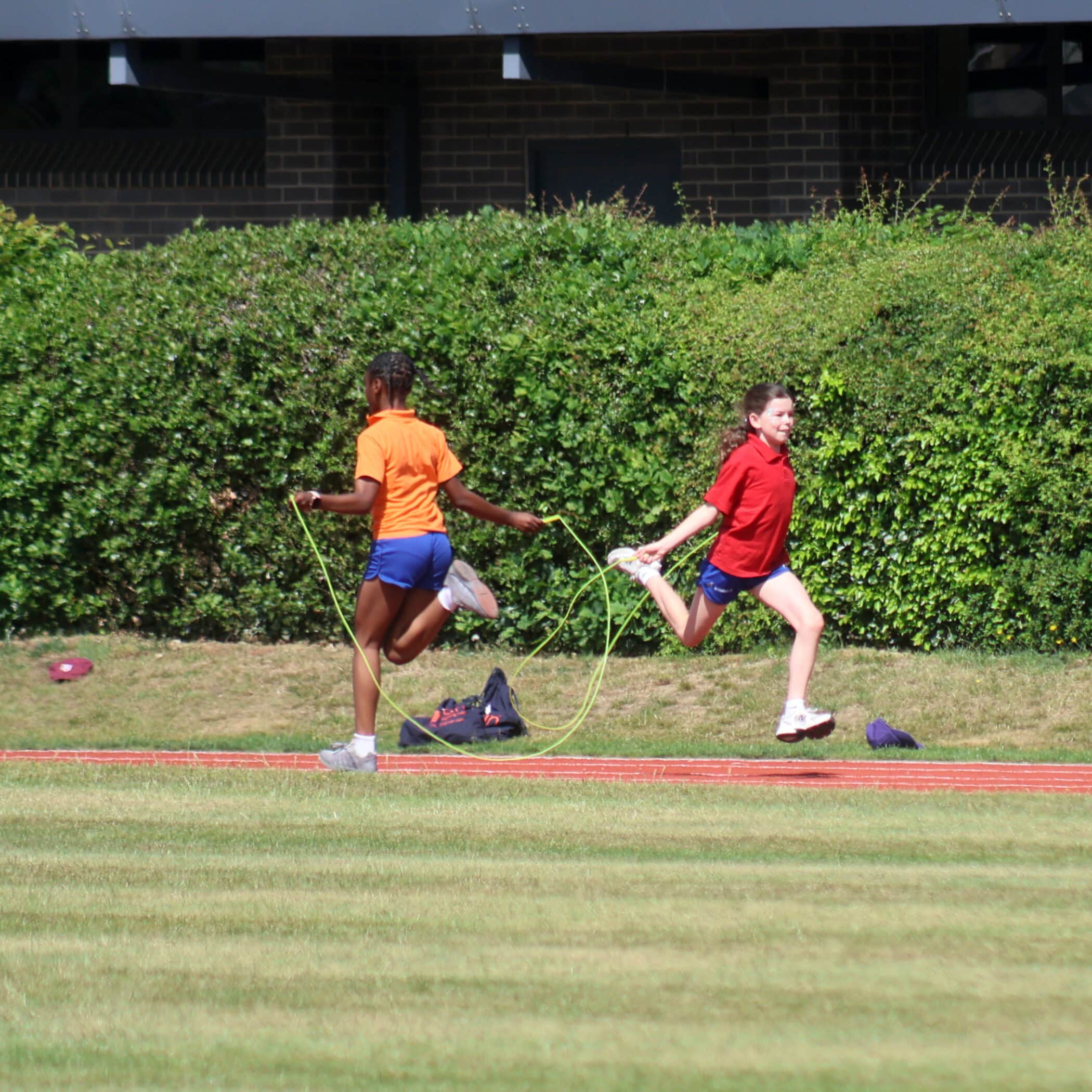 Two girls are jumping together with a skipping rope on a grassy sports field. One girl wears an orange shirt and the other wears red; both are in motion and appear to be enjoying the activity on a sunny day.