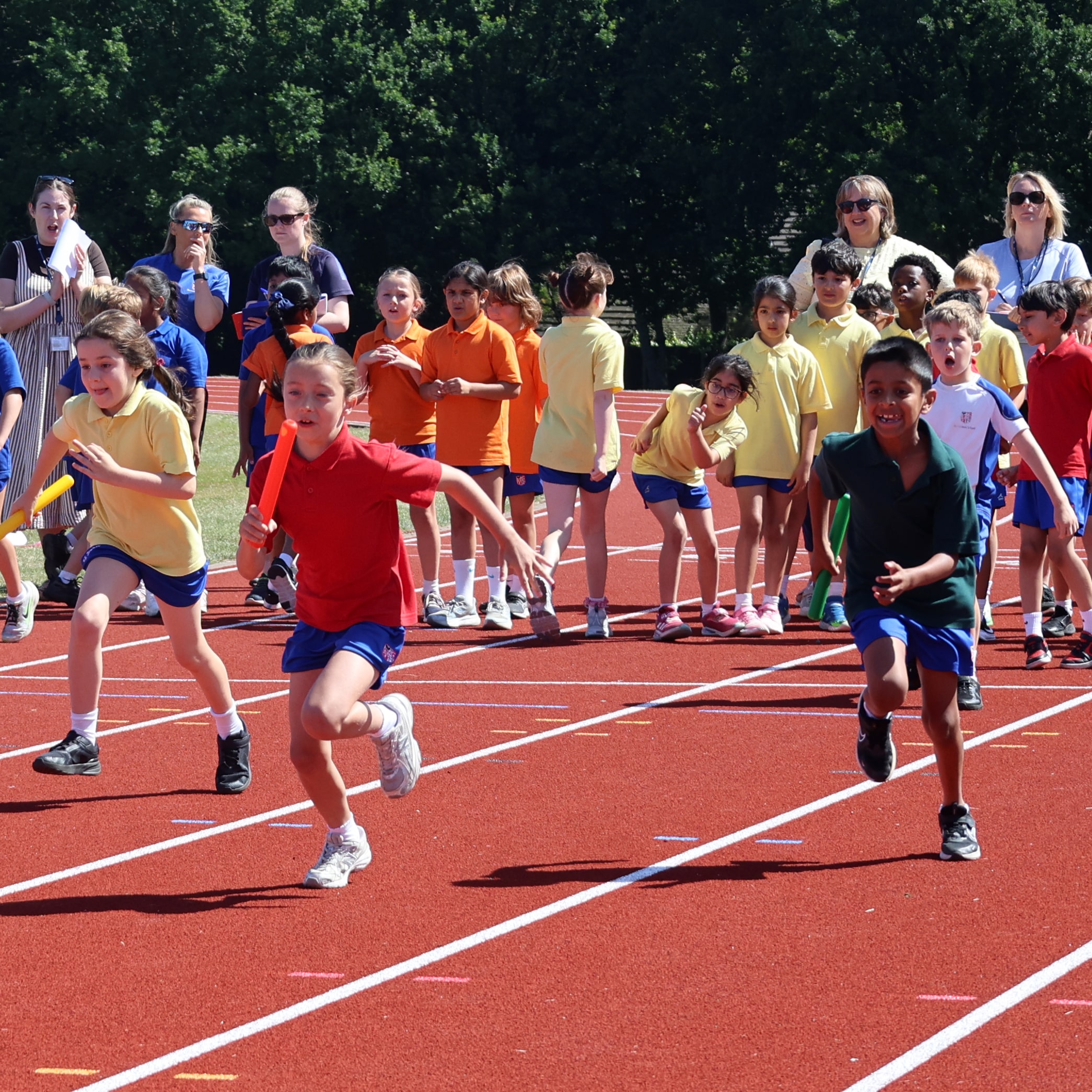 Children in colourful shirts race on a red running track during a relay event, whilst other kids and adults watch and cheer from the sidelines under sunny skies.