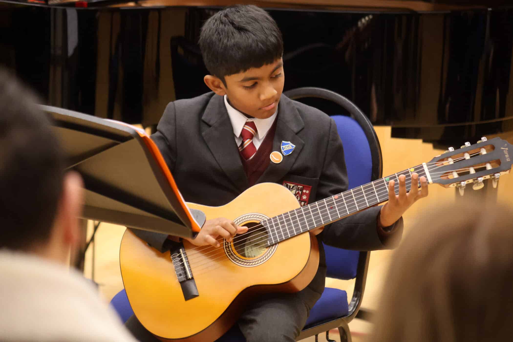 A young boy in a school uniform plays a classical guitar, reading sheet music from a stand during a performance, with an audience in the foreground, an inspiring example of why music education is important for child development.