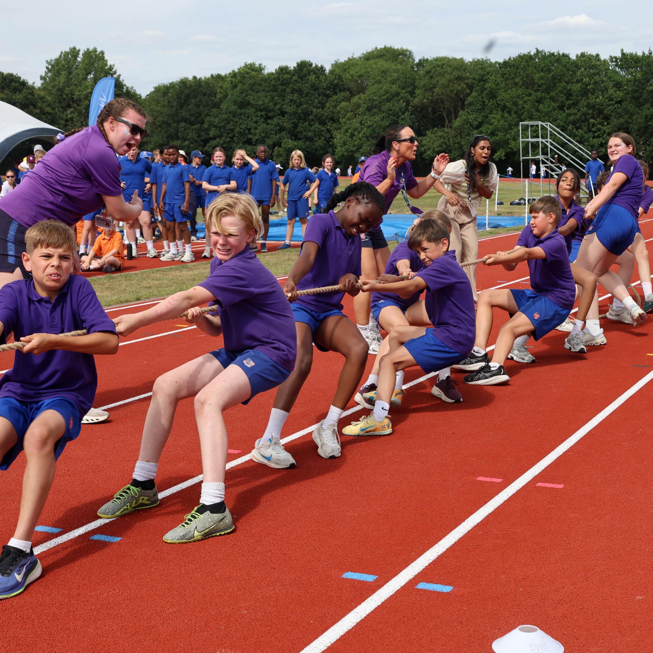 A group of children in purple shirts enthusiastically play tug-of-war on a red running track, with a crowd of people wearing blue shirts cheering in the background. Trees and outdoor sports equipment are visible.