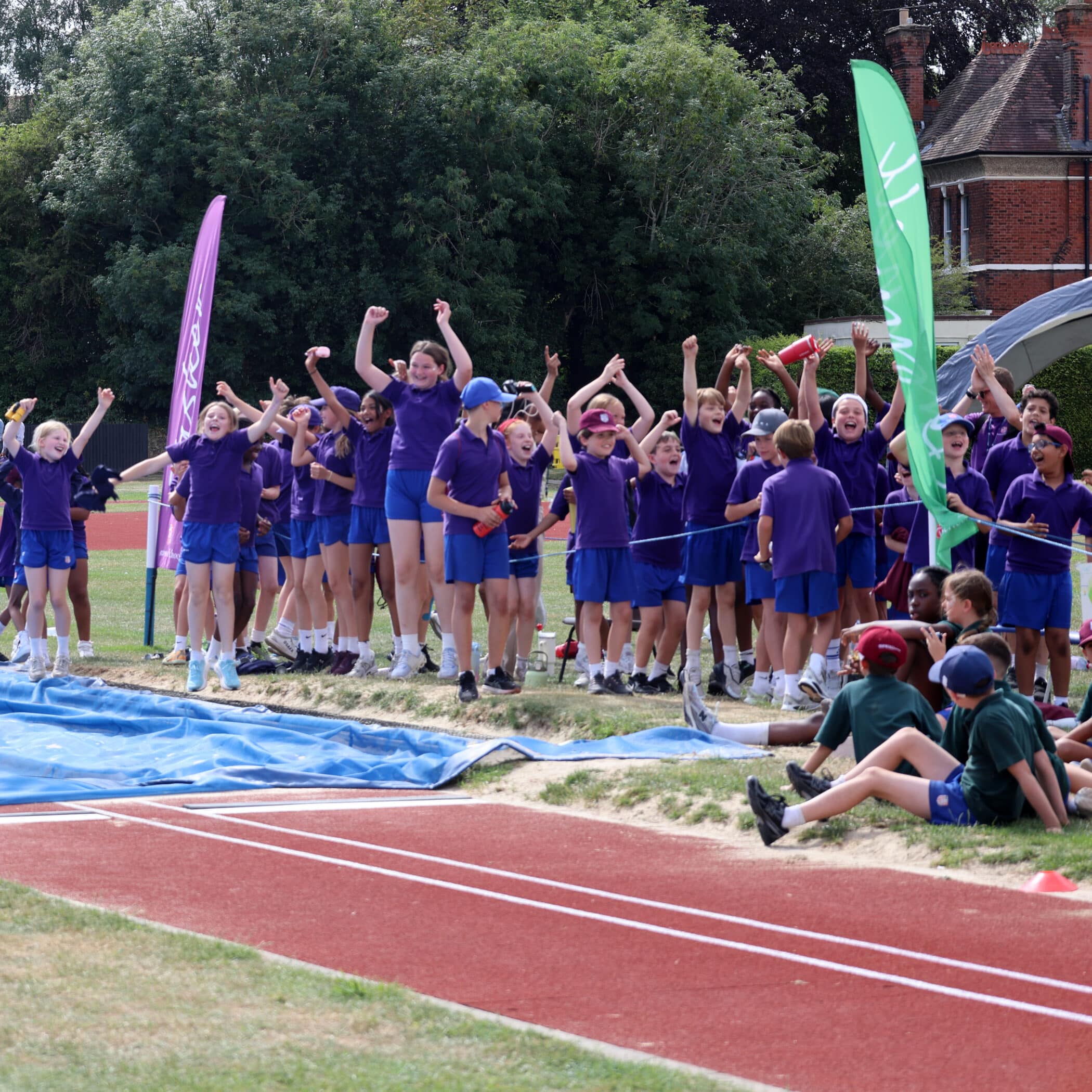 A group of children in matching purple sports kits cheer and celebrate beside a long jump pit at an athletics track, while a few others in green kits sit on the grass nearby.