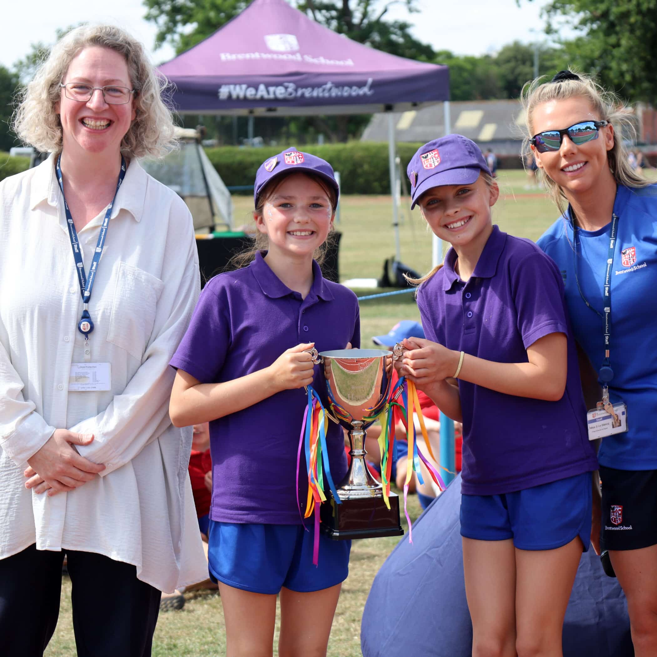 Two girls in purple sports kits hold a trophy with colourful ribbons, smiling. Two women stand beside them, also smiling. A marquee with Brentwood School text is visible in the background on a sunny day.