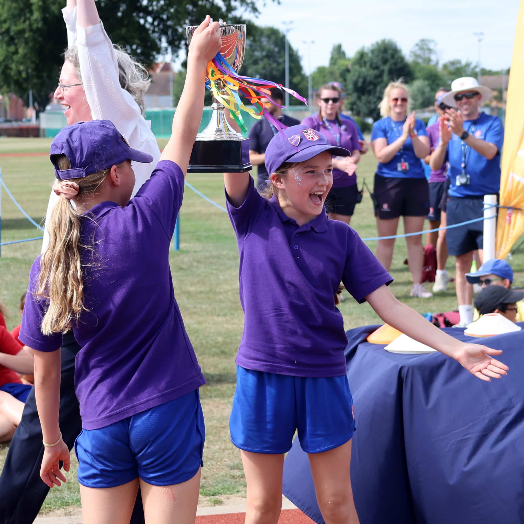 Two girls in purple shirts and caps hold up a trophy with colourful ribbons, smiling joyfully. People in the background clap and celebrate outdoors on a sunny day, likely at a school sports event.