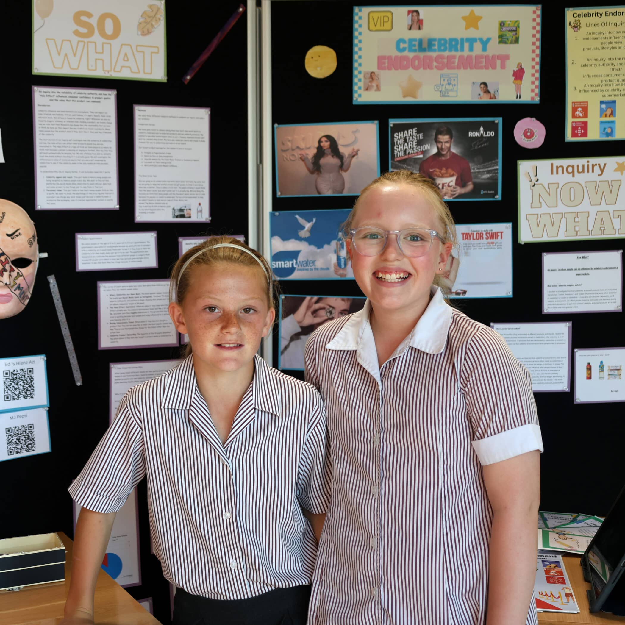 Two girls in striped school uniforms stand smiling in front of a display board about celebrity endorsement, featuring posters, text, and images related to marketing and advertising.