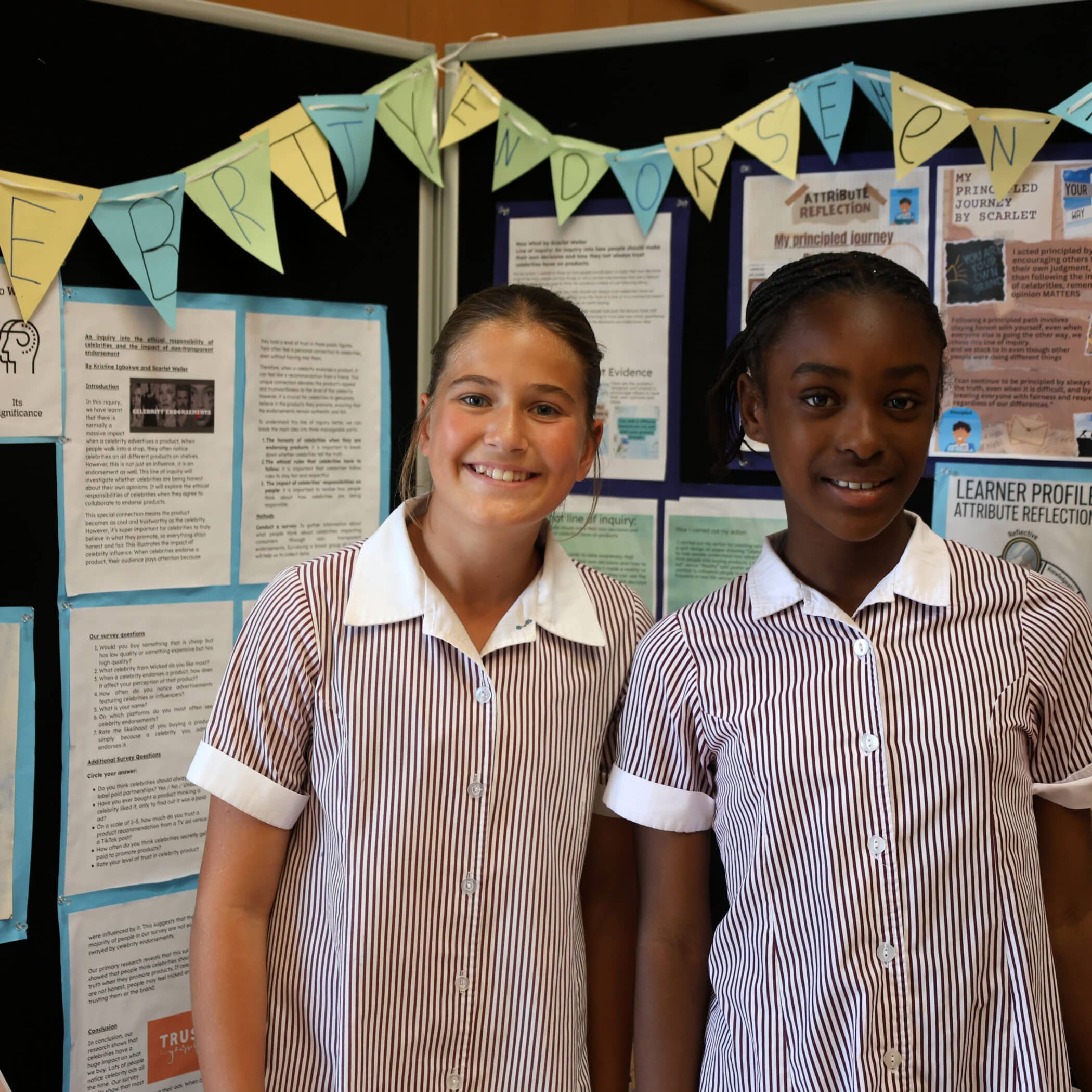 Two schoolgirls in striped uniforms smile in front of a display board covered with posters and pupils' work. A banner with cut-out letters hangs above them.