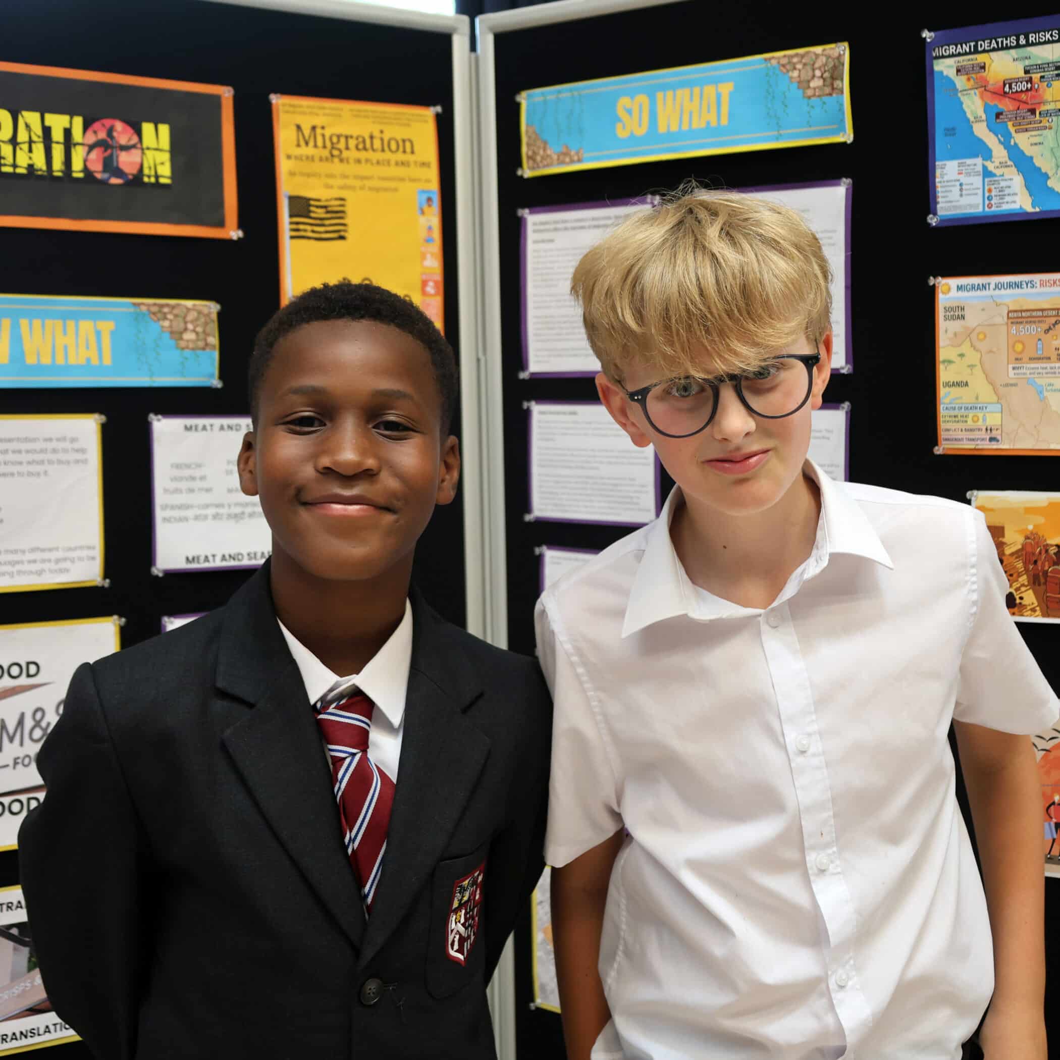 Two boys stand in front of a school display about migration. One wears a suit and tie; the other wears glasses and a white shirt. Colourful posters and maps with migration information are visible behind them.