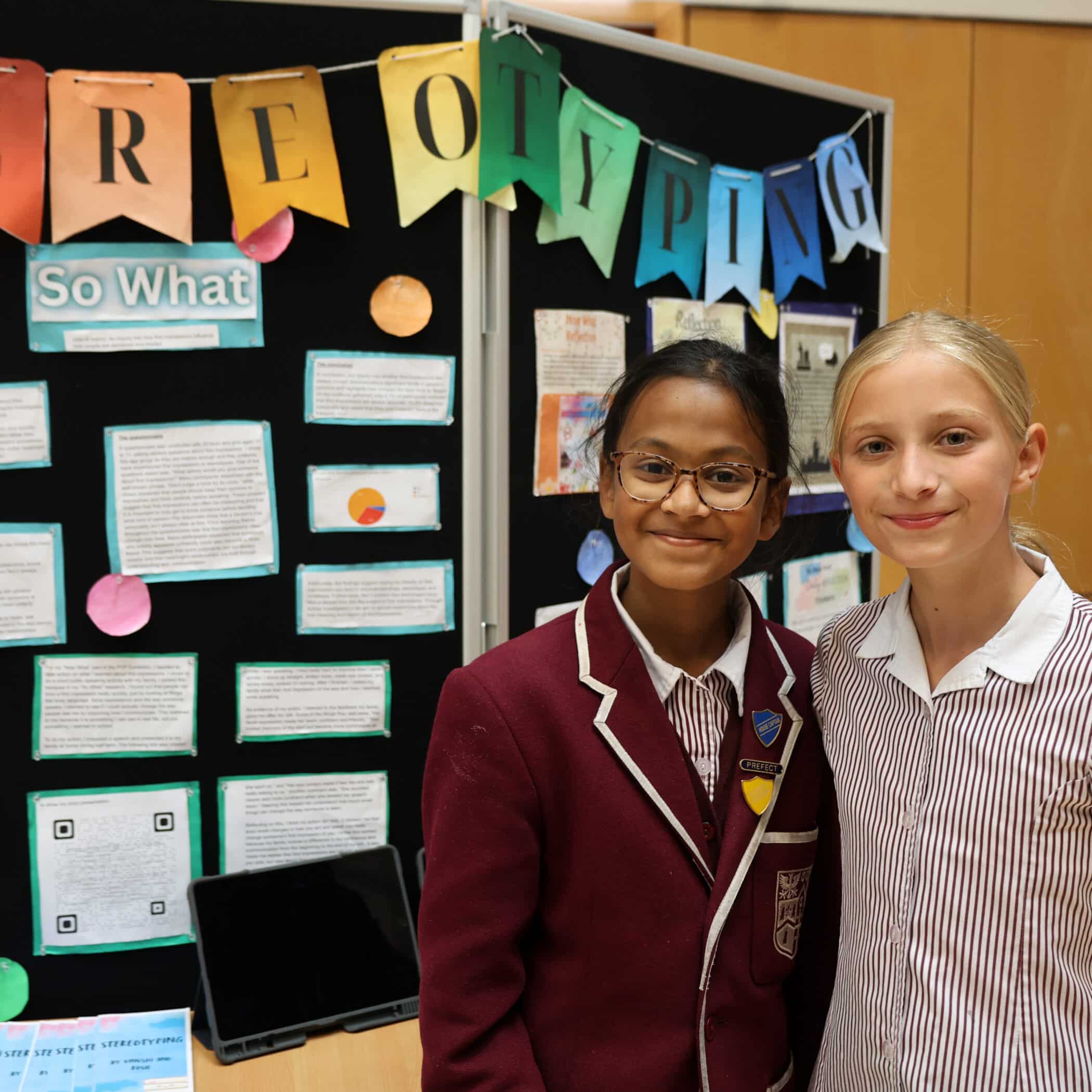 Two girls stand smiling in front of a school project display titled “Stereotyping.” One wears a striped shirt and the other a maroon blazer and glasses. The display includes colourful banners, text, and printed materials.