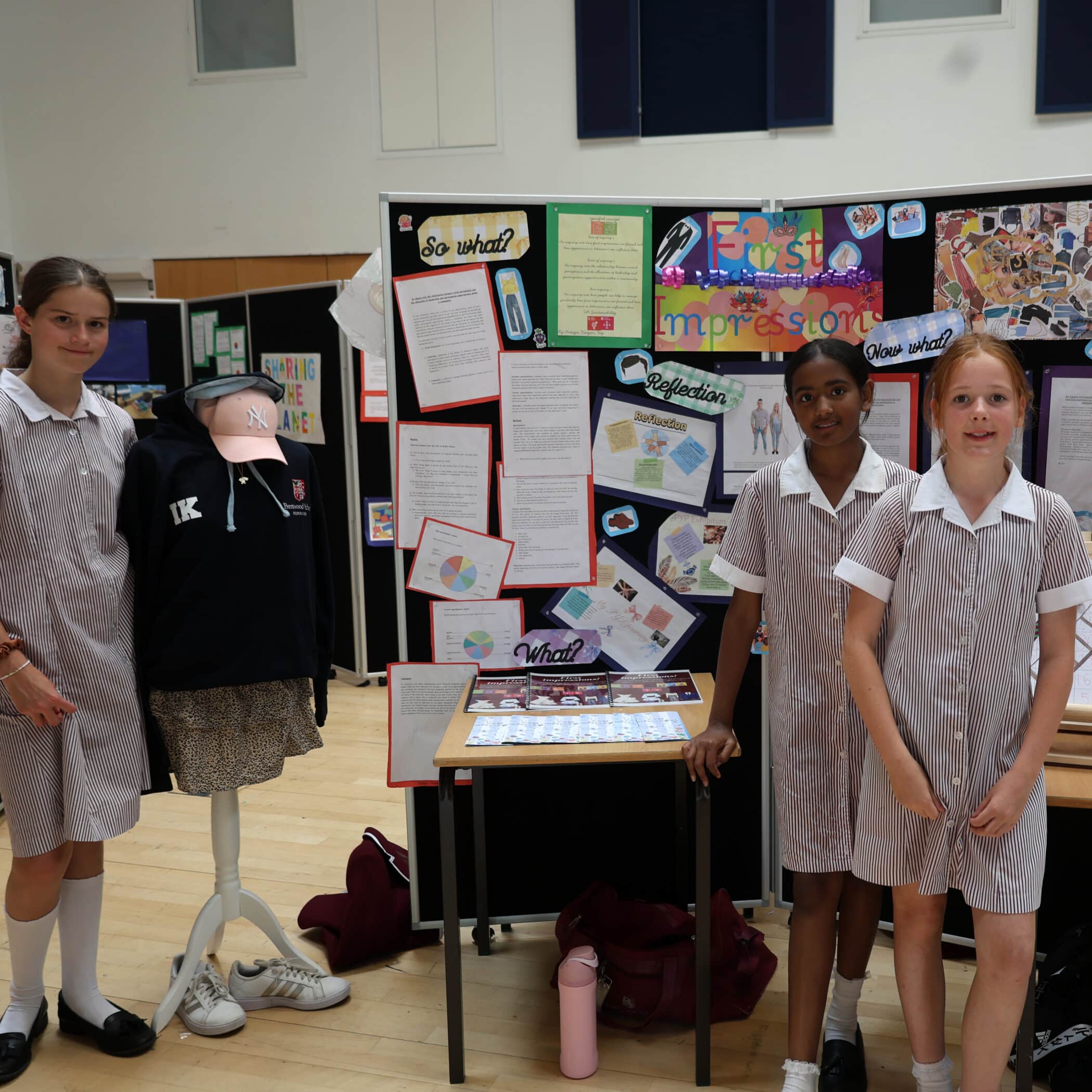 Three schoolgirls in striped uniforms stand in front of a colourful display board titled First Impressions, featuring posters, drawings, and a mannequin dressed in a school top and skirt, inside a classroom setting.