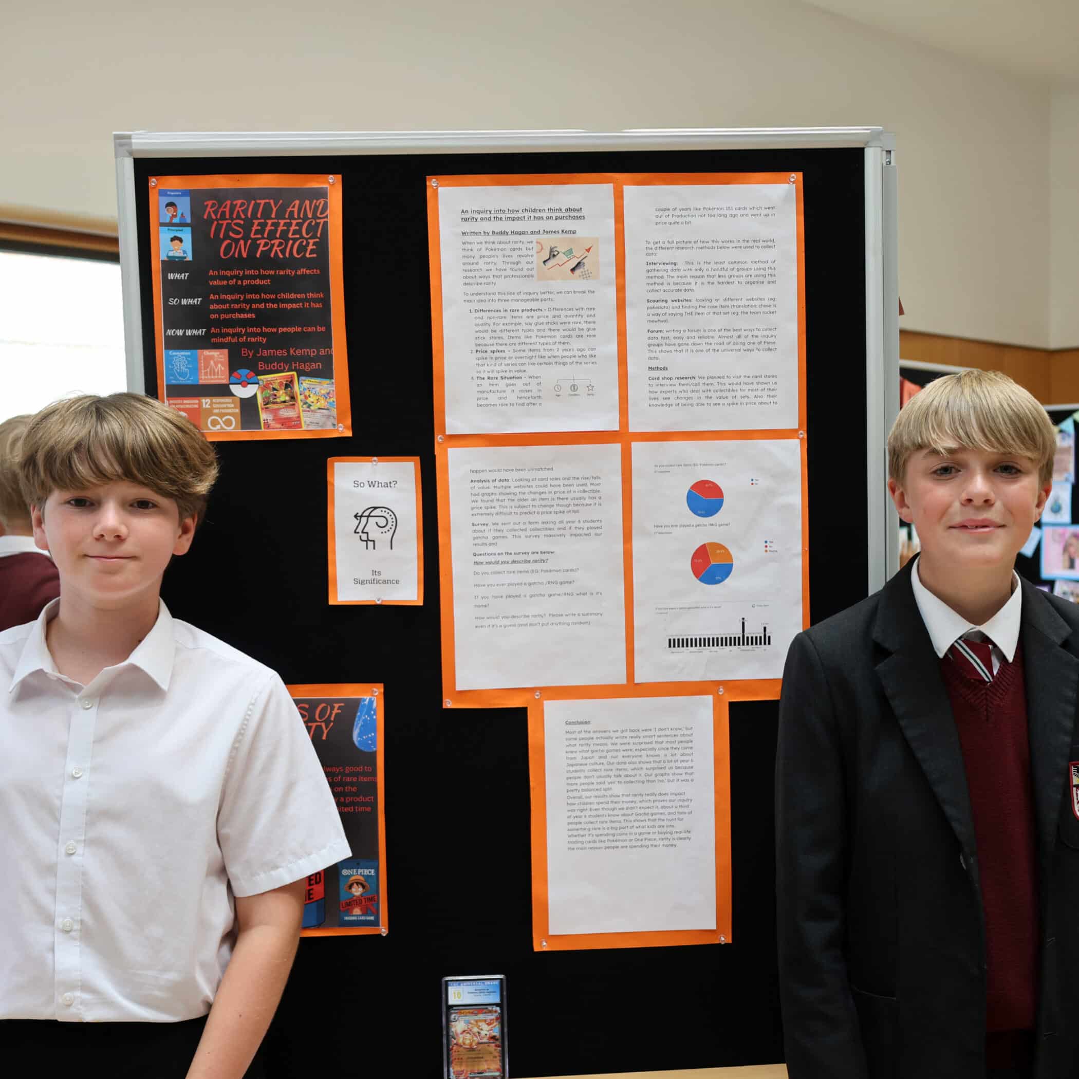 Two boys stand in front of a science fair project display board titled Rarity and Its Effect on Price, featuring charts, graphs, and text on orange-bordered papers. Both are wearing school uniforms.