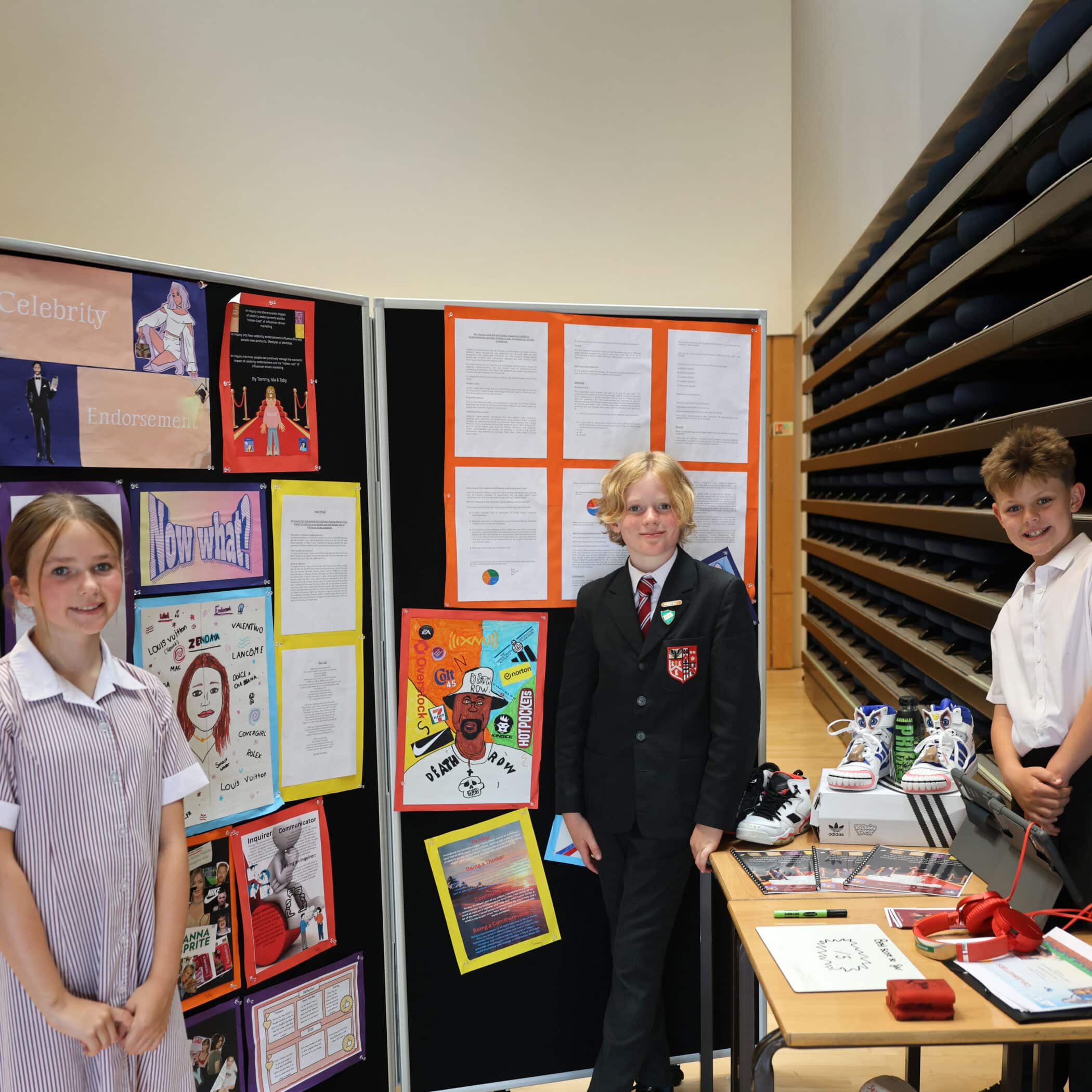 Three schoolchildren stand by a display board with colourful posters and written projects. The board features artwork, written reports, and photos. A table in front holds shoes and other small items. They are in a large, bright room.