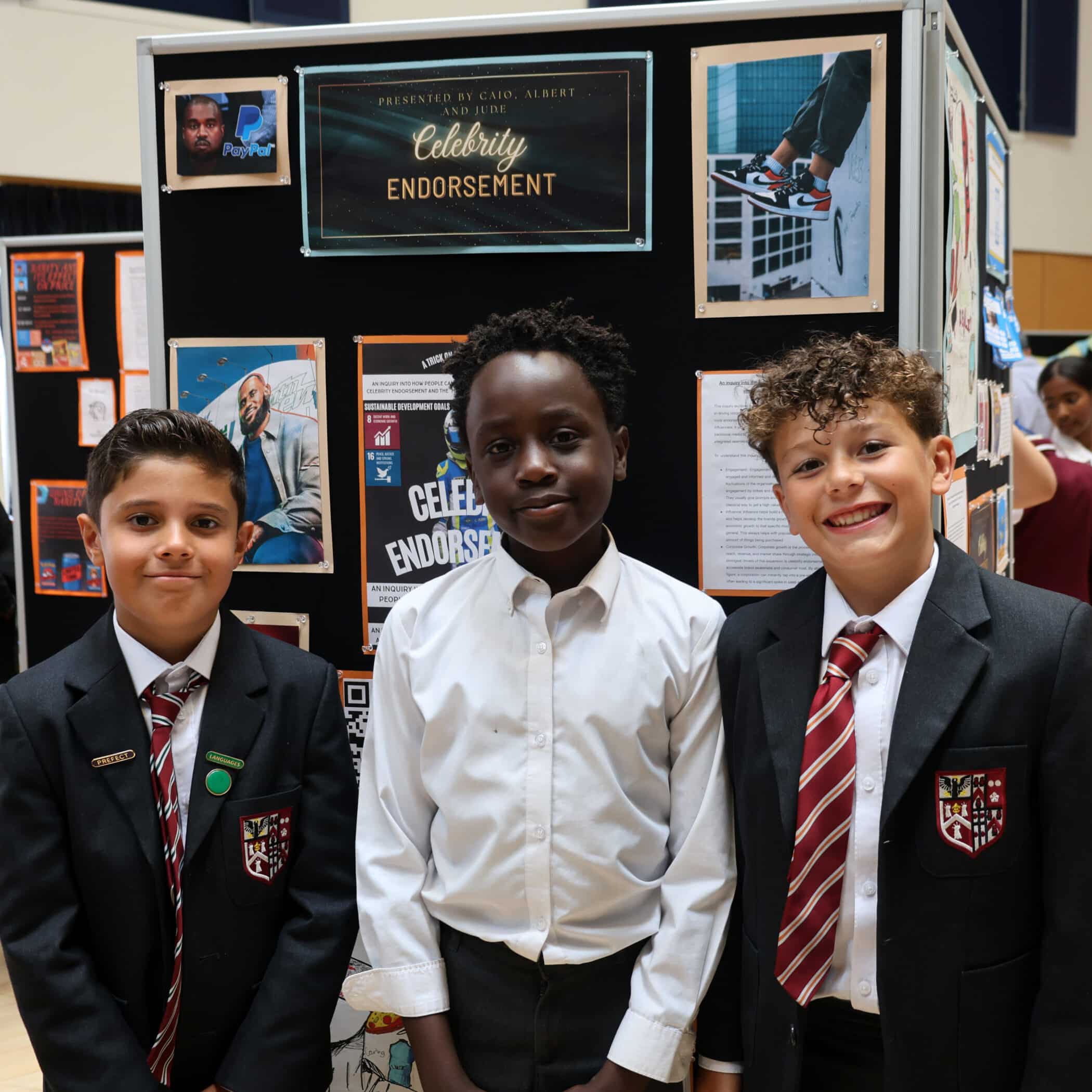 Three boys in school uniforms stand in front of a presentation board titled Celebrity Endorsement. The board displays various images and information about celebrities and advertising.