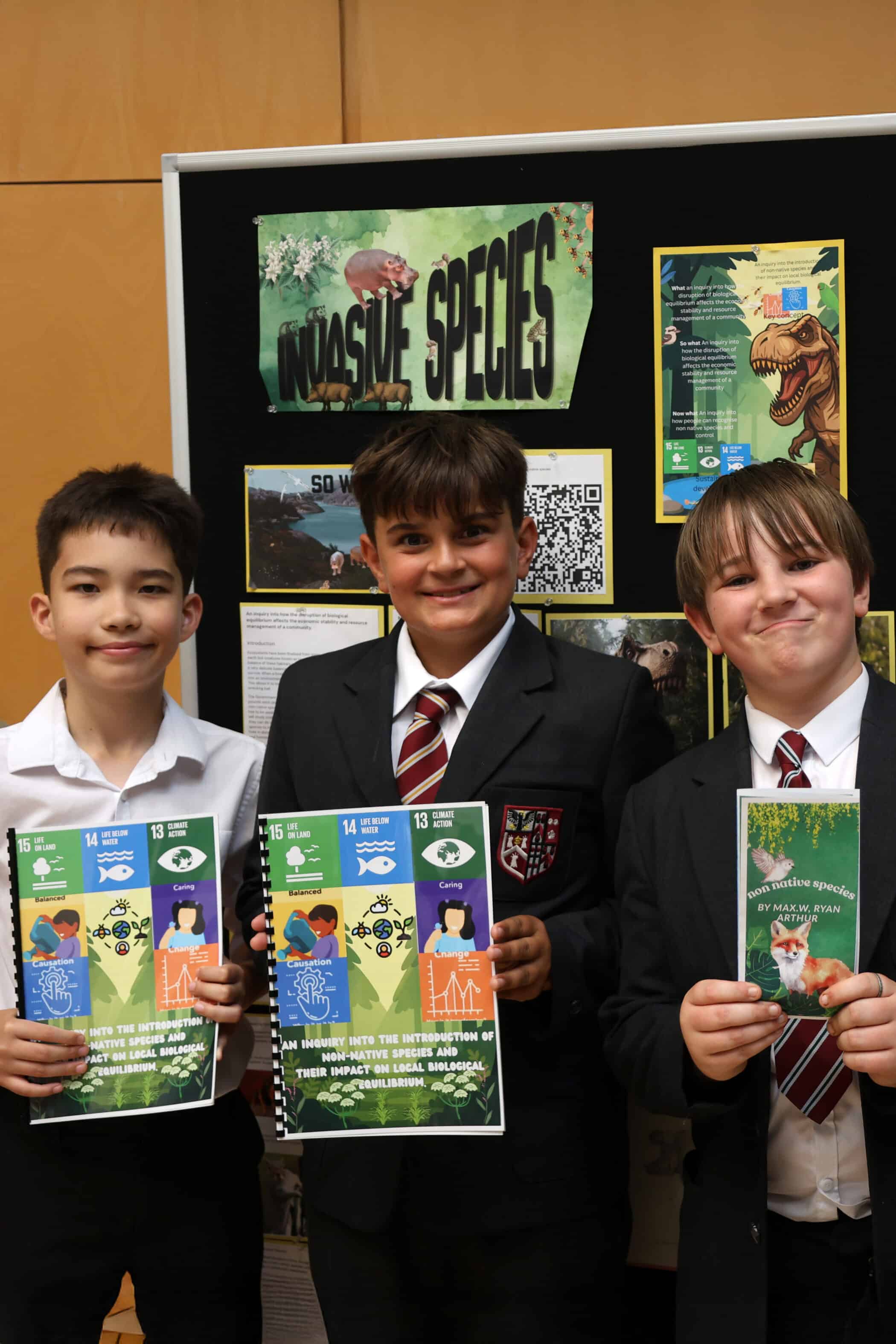 Three boys in school uniforms stand smiling in front of a display board about invasive species, each holding informative leaflets. The board features posters, images of animals, and a large Invasive Species sign.
