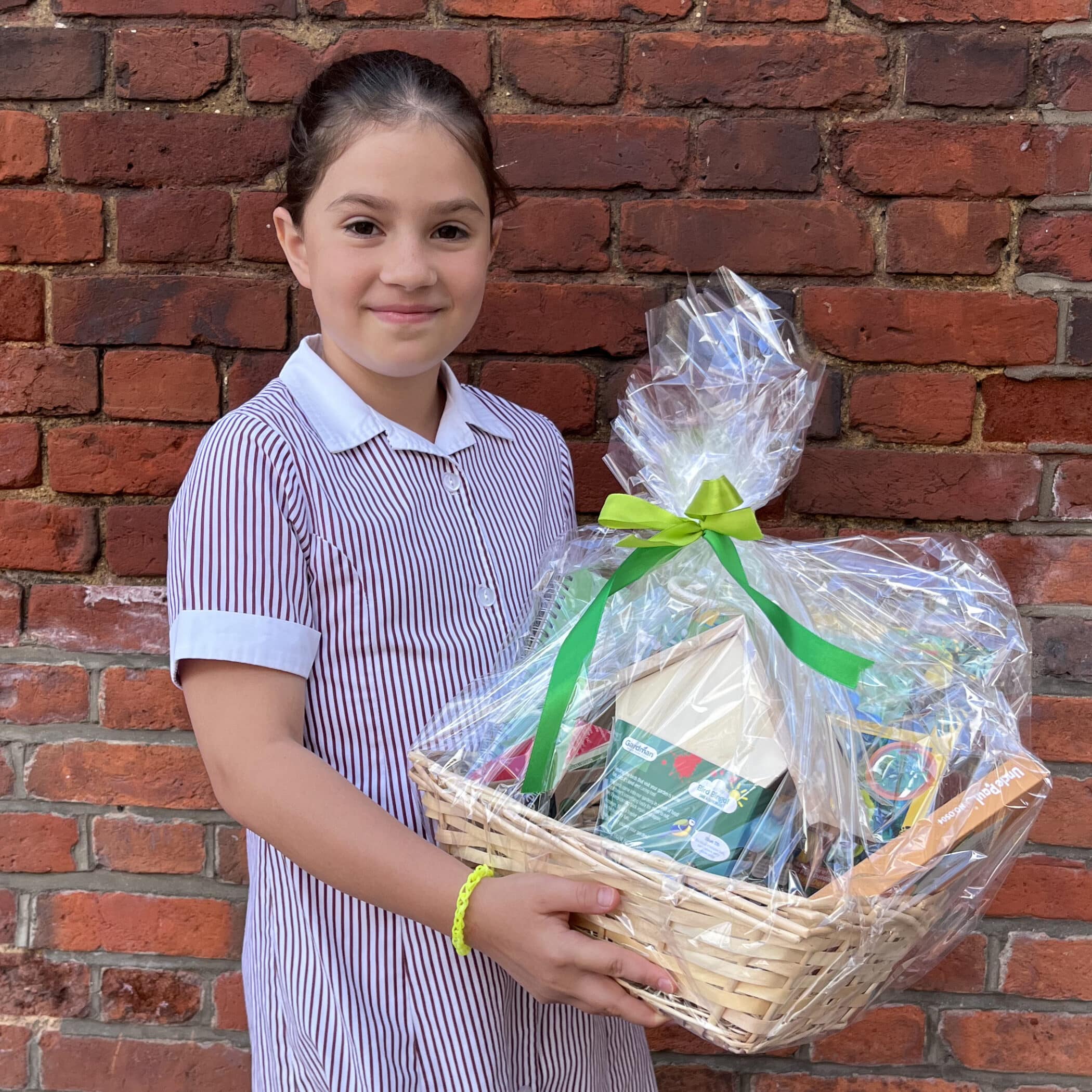 A young girl in a striped school uniform holds a gift hamper wrapped in clear plastic with a green ribbon, standing in front of a red brick wall.