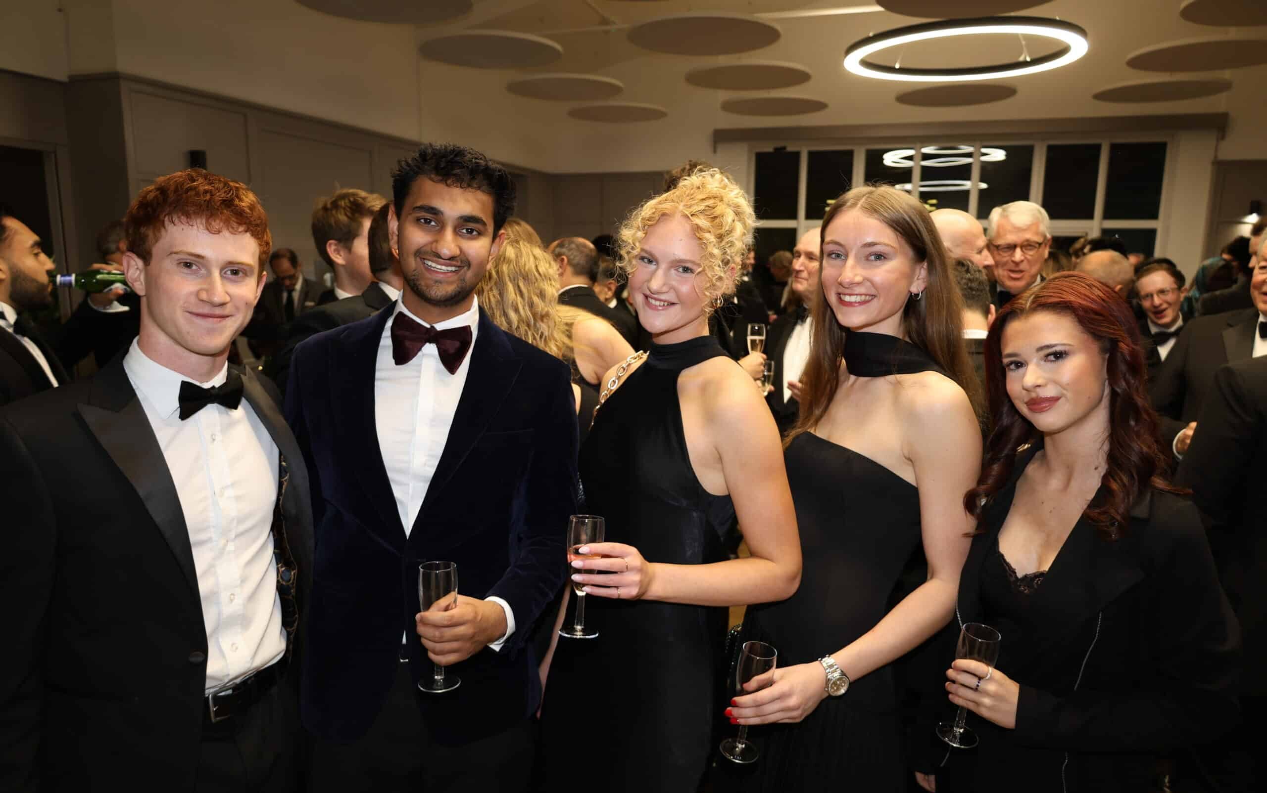 Five people dressed formally stand together at a party hosted by the Brentwood School Association, smiling and holding champagne glasses, with other guests mingling in the background under modern circular lights.