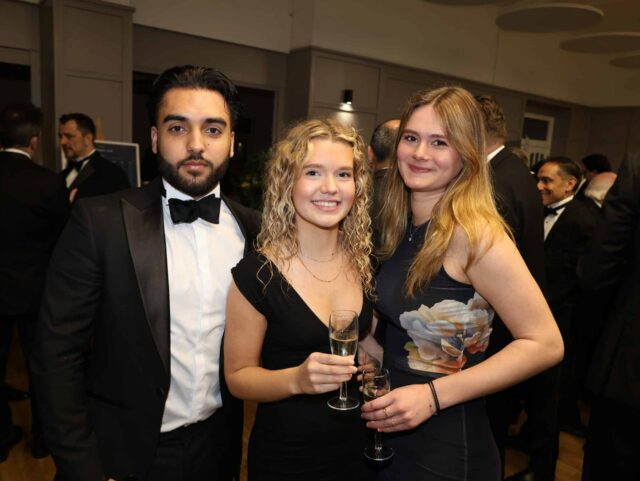 Three people dressed formally stand together at an event organised by the Brentwood School Association, smiling at the camera. The two women hold glasses of champagne. Other guests in suits are visible in the background.