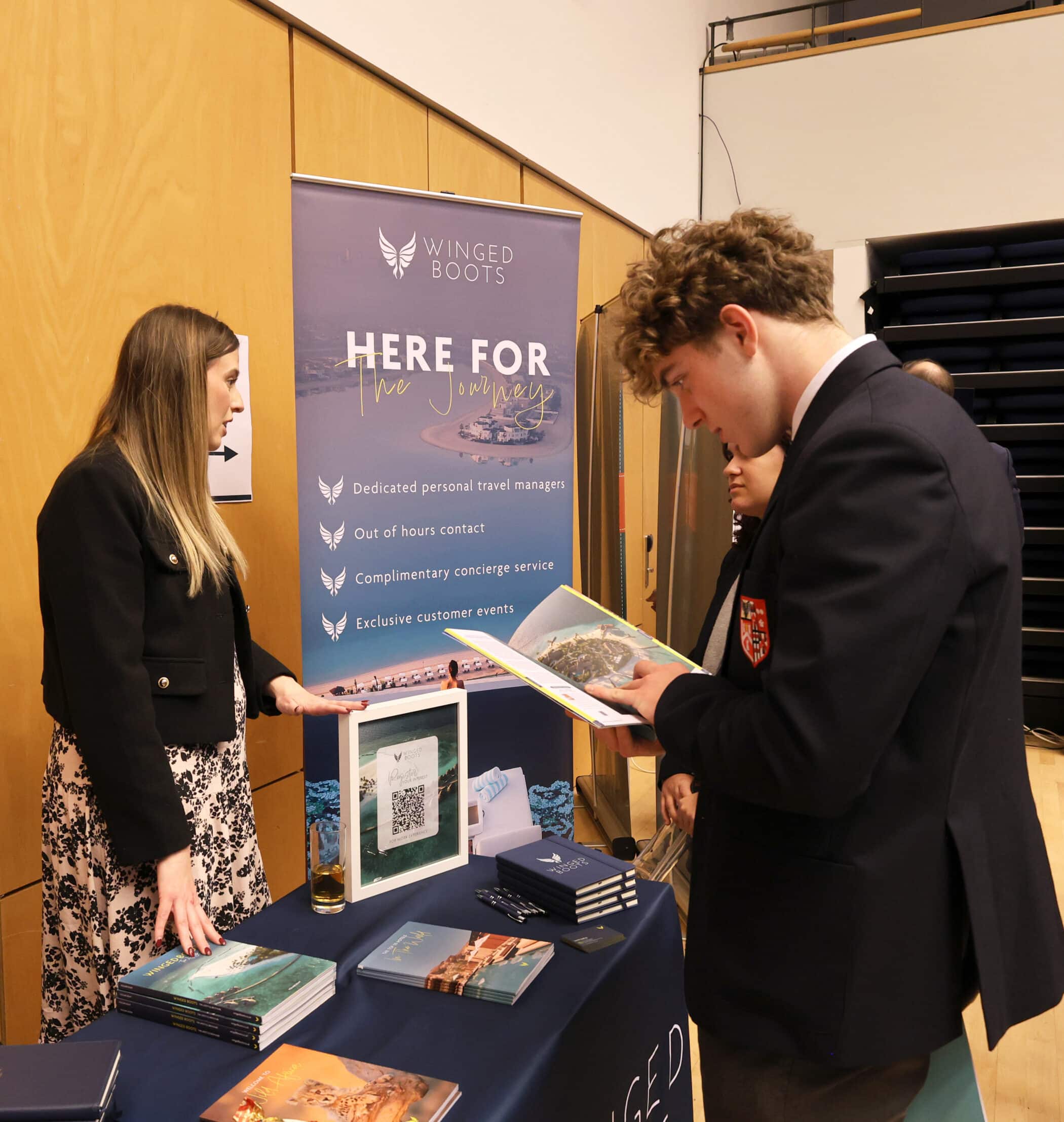 A woman stands behind a Winged Boots travel company stand while a young man in a suit, attending the Brentwood School Careers Convention, looks at a brochure. The stand displays brochures, a QR code, and promotional banners.