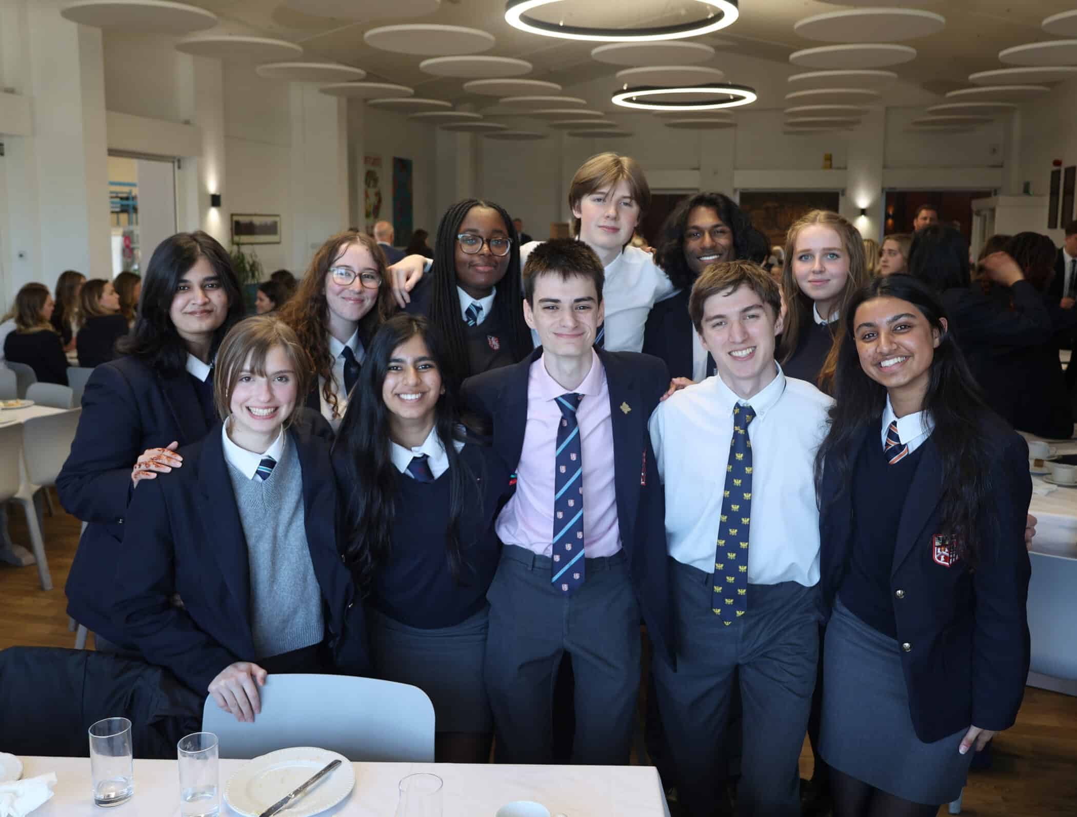A group of students in school uniforms smile and pose together in the Brentwood School Dining Halls, some standing and some sitting at a table with empty plates and glasses. Round ceiling lights are visible above them, capturing a lively moment of camaraderie among Brentwood School Association members.