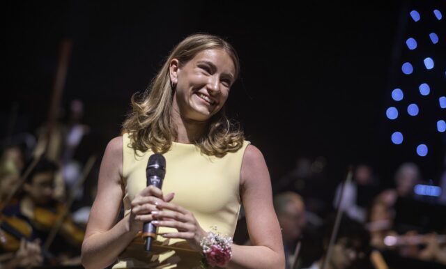 A young woman in a light yellow dress smiles whilst holding a microphone on stage, with an orchestra playing in the background. Blue lights glow on the right, celebrating at the Brentwood School prom concert.