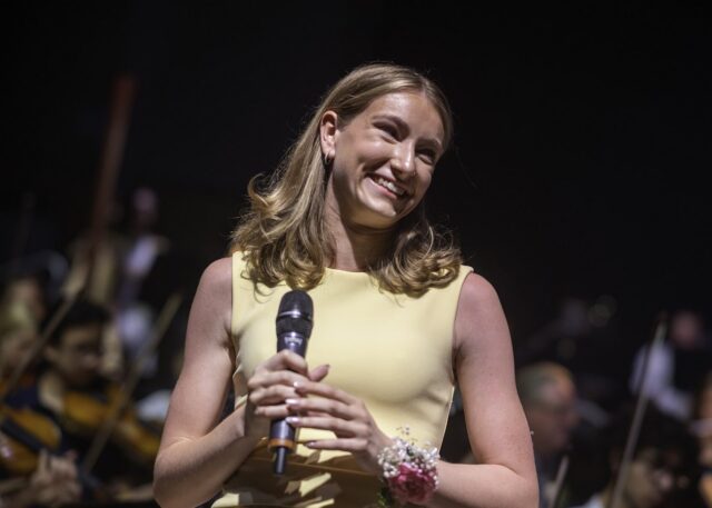 A young woman in a light yellow dress smiles whilst holding a microphone on stage, with an orchestra playing in the background. Blue lights glow on the right, celebrating at the Brentwood School prom concert.