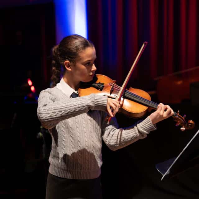 A young person wearing a grey jumper plays the violin on stage, focused on sheet music, with red curtains and stage lights in the background, performing as part of a concert.