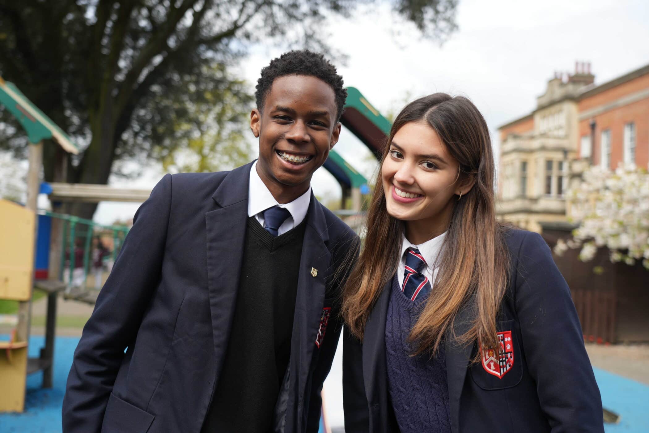 Two students in school uniforms stand outside, smiling at the camera. They are wearing navy blazers and ties, representing the opportunities provided by bursaries to attend the school. Behind them, trees and a school building are visible in the background.
