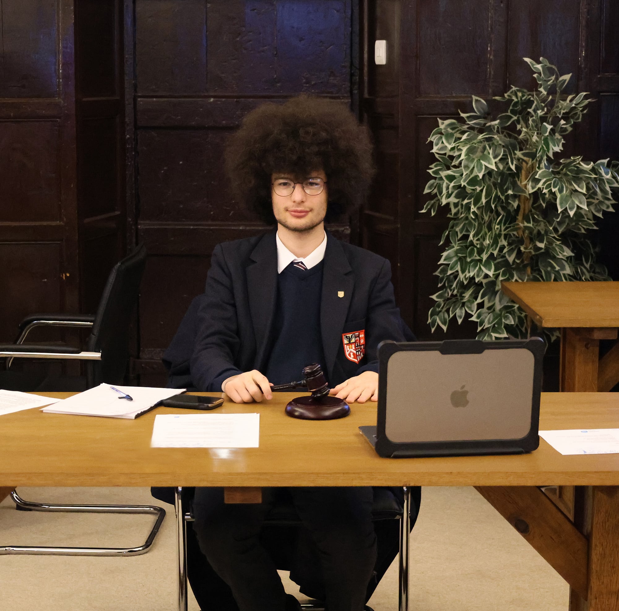 A young person with curly hair and glasses, dressed in a suit, sits at a wooden desk with papers, a gavel, and a laptop. They are in a formal room with dark panelling and a potted plant in the background, suggesting an official setting, perhaps during a Brentwood School Foundation meeting or event.