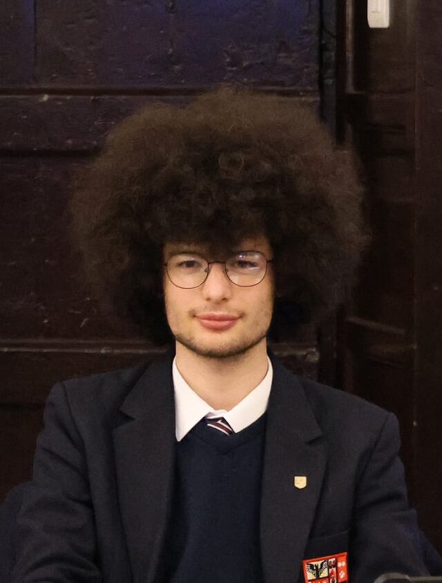 A person with curly hair in a suit sits at a desk in a formal room with dark wood panelling.