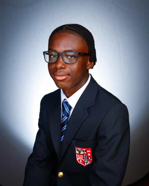 A young person wearing glasses and a dark suit with a striped tie sits in front of a white background, looking at the camera with a slight smile. A crest, representing Brentwood School is visible on the blazer pocket.