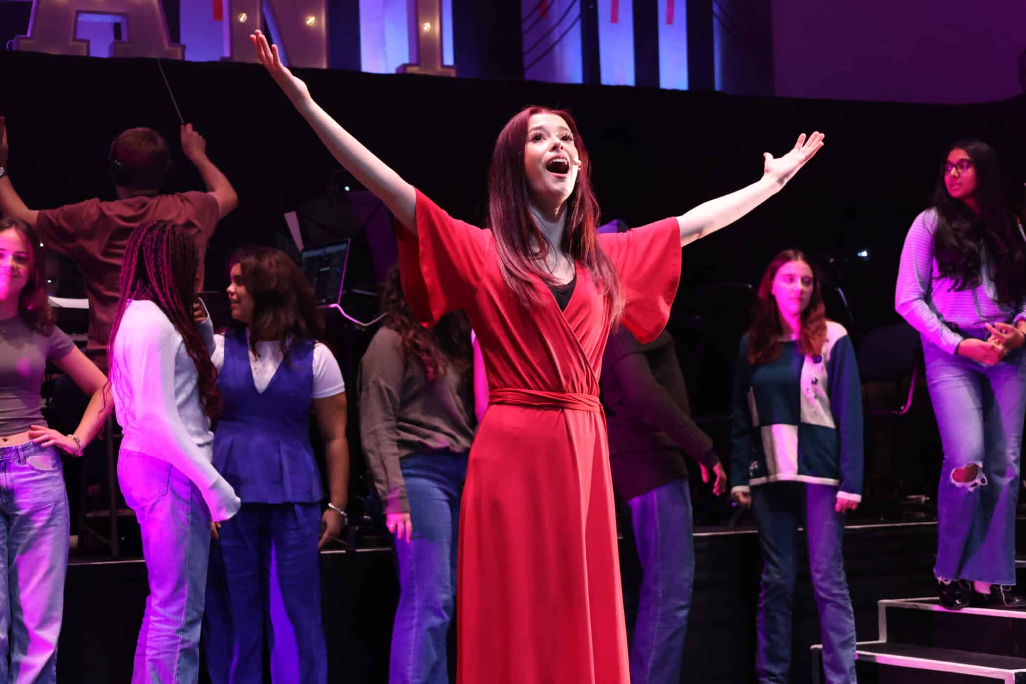 A woman in a red dress stands centre stage with arms outstretched, singing passionately. Several people stand behind her on stage, illuminated by colourful lights.