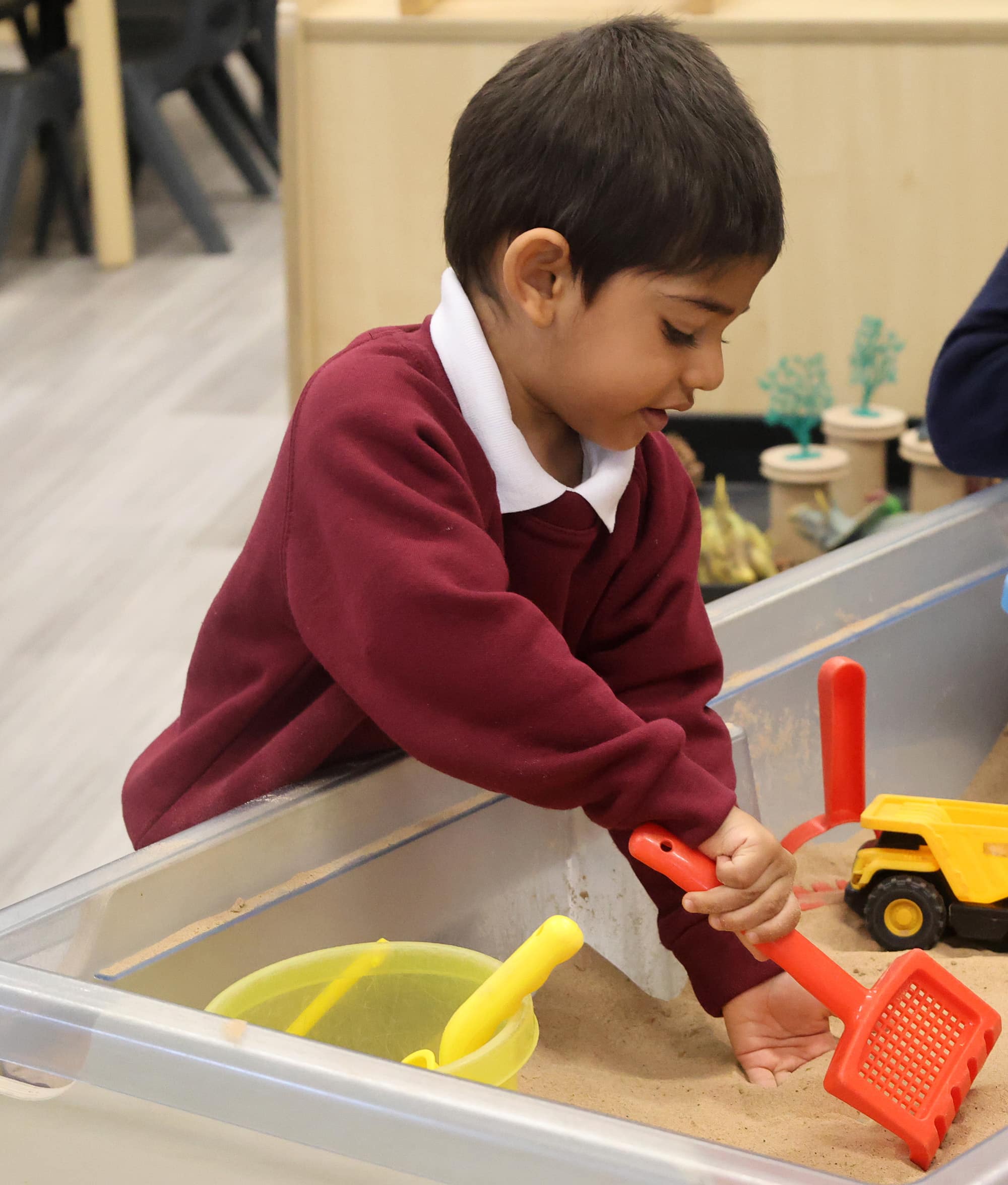 A young child in a maroon jumper plays at a sand table, using a red spade to dig near a yellow bucket and toy dumper truck, enjoying creative playtime in Foundation.