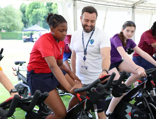 A group of children on stationary bikes in a marquee are guided by a smiling staff member. The children are raising funds for the Brentwood School Foundation and wear sports uniforms and appear focused on the exercise activity.
