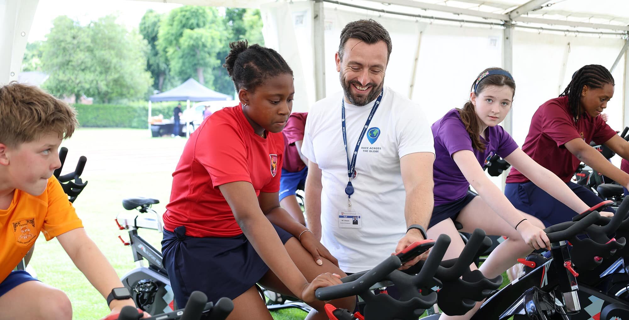 A group of students ride stationary bikes in a marquee. All appear focused, wearing colourful shirts and shorts, participating in an organised fitness activity sponsored by the Brentwood School Foundation.