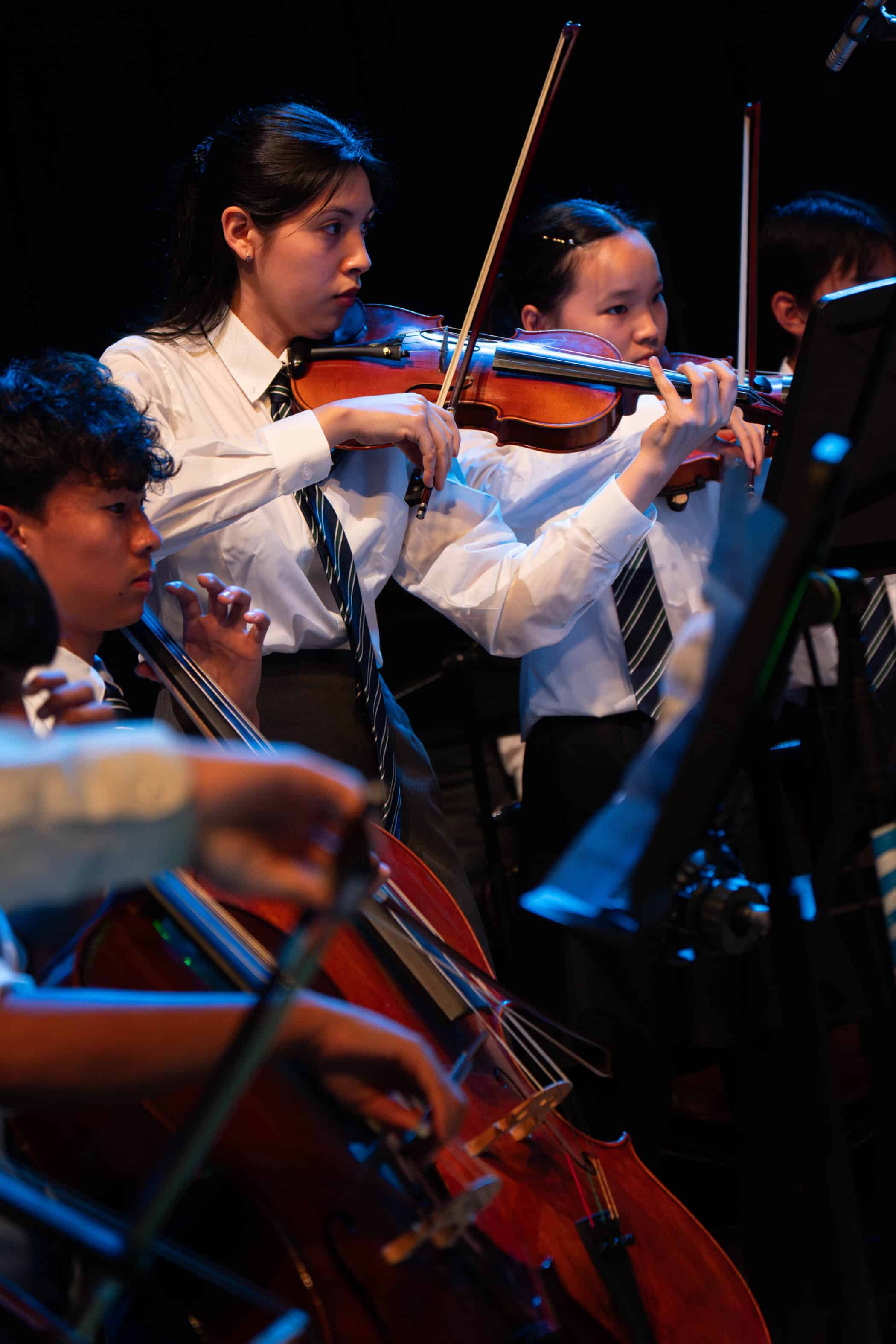 Students in white shirts and ties play violins and cello in an orchestra, focused on their performance under stage lighting against a dark background, a showcase of the remarkable talent nurtured by the Specialist Music Programme.