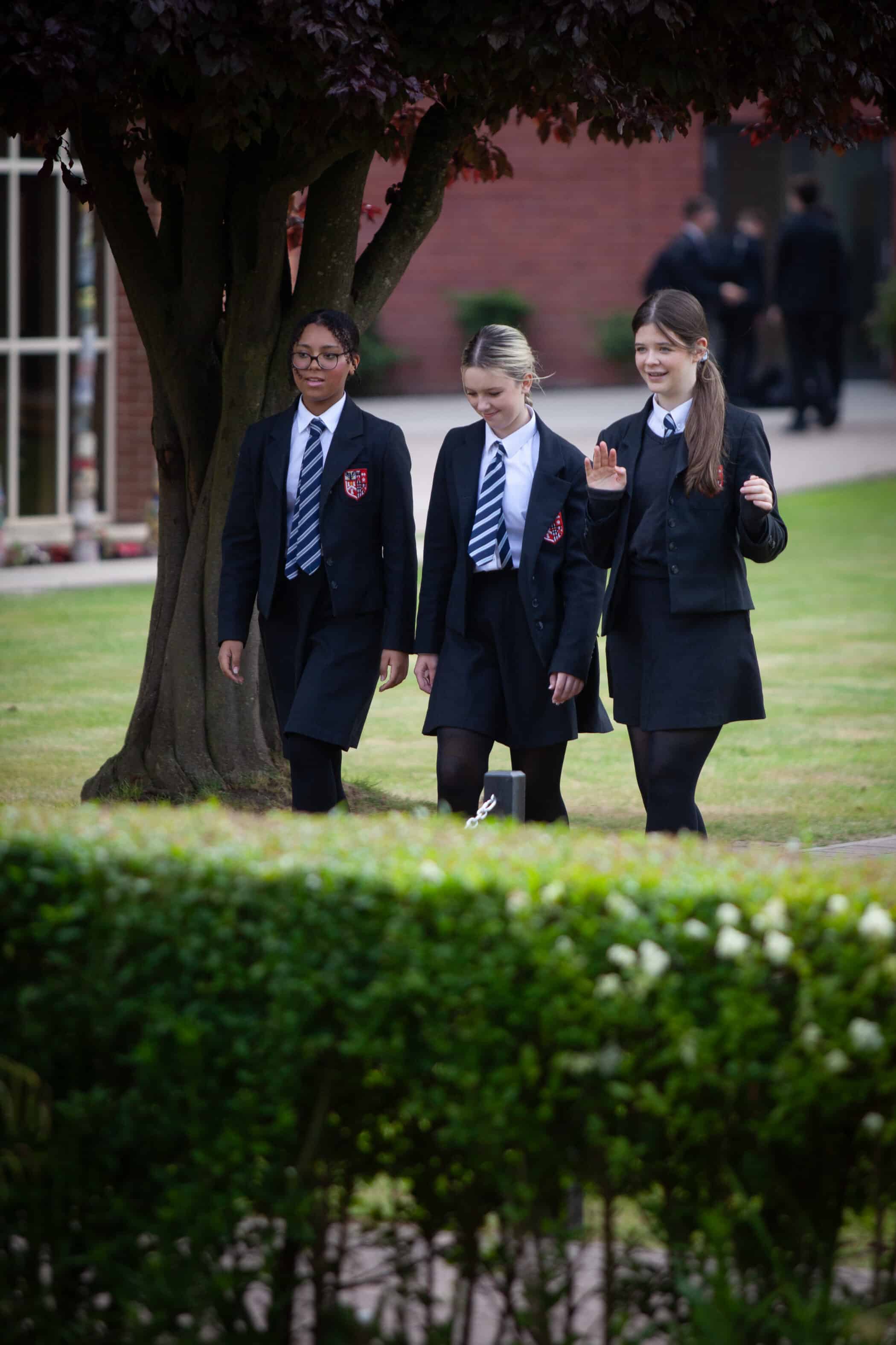 Three students in school uniforms walk together on a grassy area, talking and smiling, near a tree and a hedge, with school buildings and coach routes visible in the background.