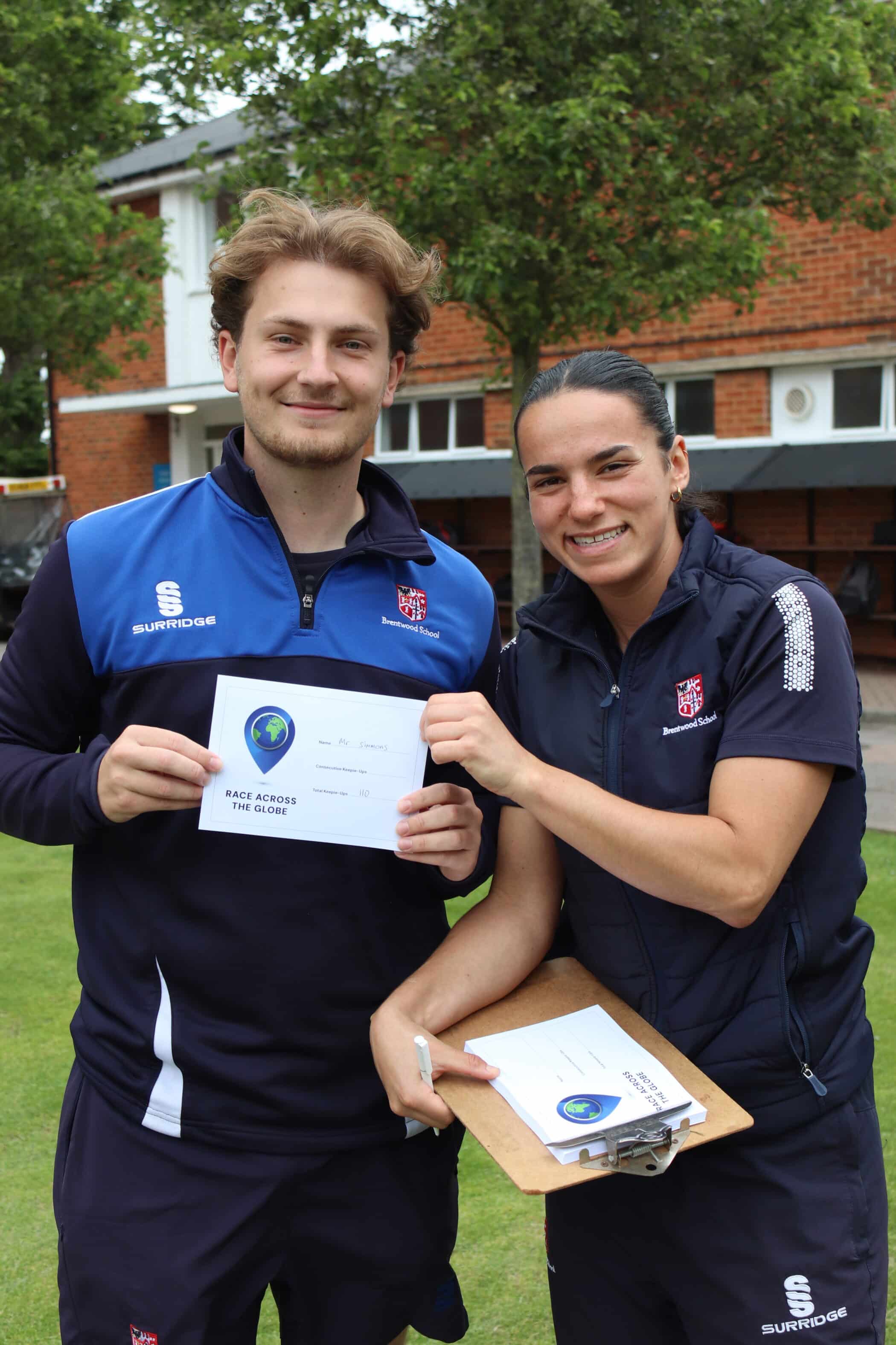 Two people in sportswear smile outdoors while holding a Brentwood School Foundation certificate together. The person on the right also holds a clipboard. Trees and a brick building are in the background.