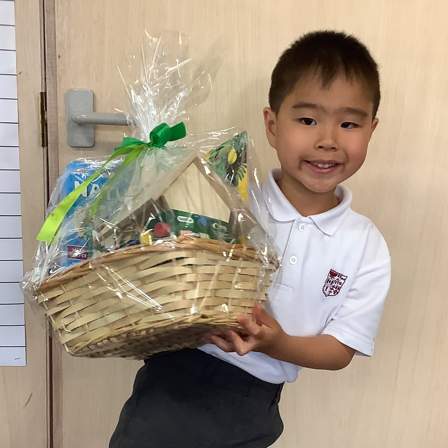 A young boy in a white polo shirt smiles whilst holding a large wicker gift hamper wrapped in clear plastic with a green ribbon, standing indoors in front of a wooden door.