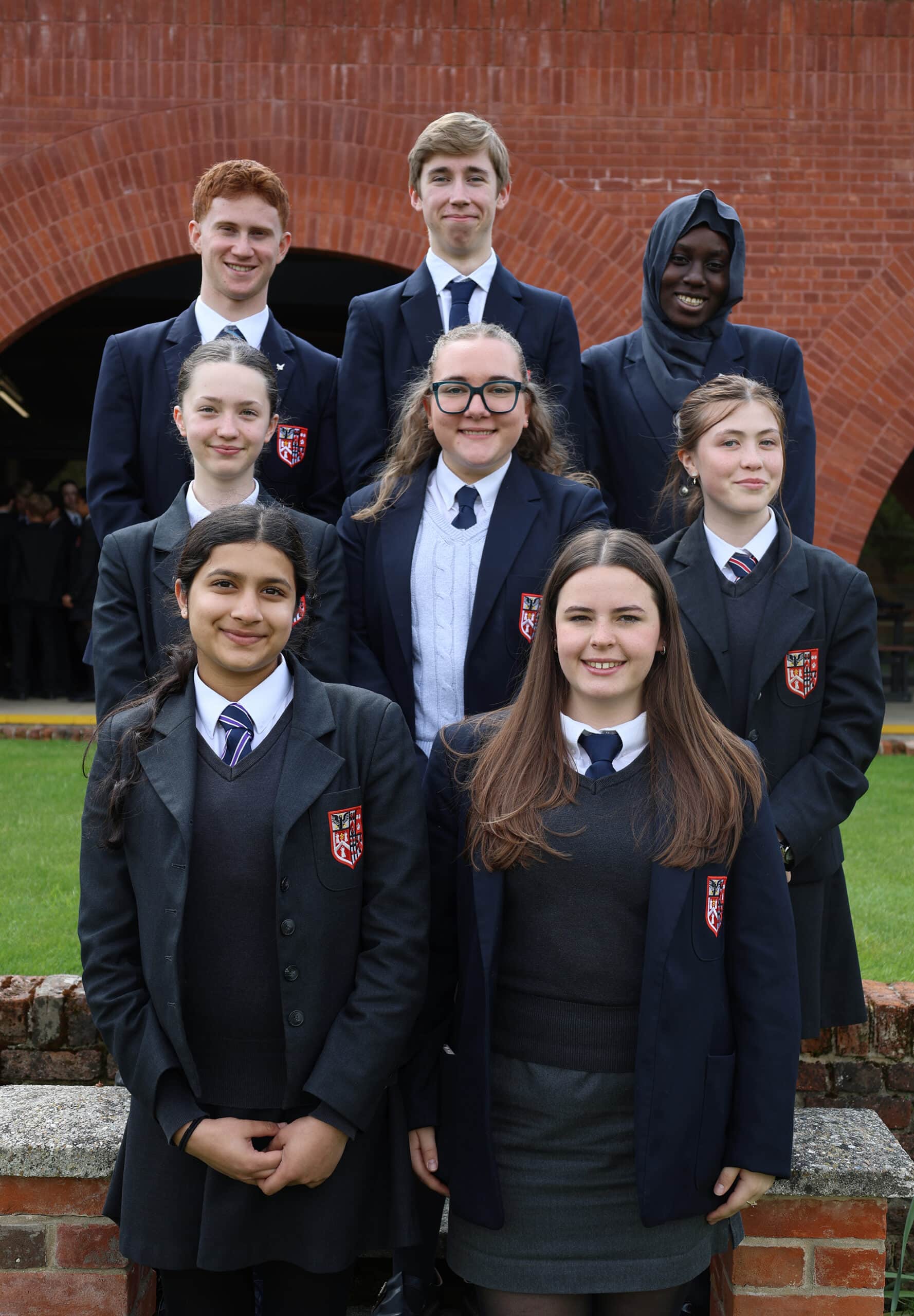 Eight students in school uniforms stand outside in front of a brick building, smiling for a group photo. Arranged in three rows with a grassy lawn behind them, the group reflects the strong sense of community often found in Sixth Forms, where students build friendships and develop skills that prepare them for university life.