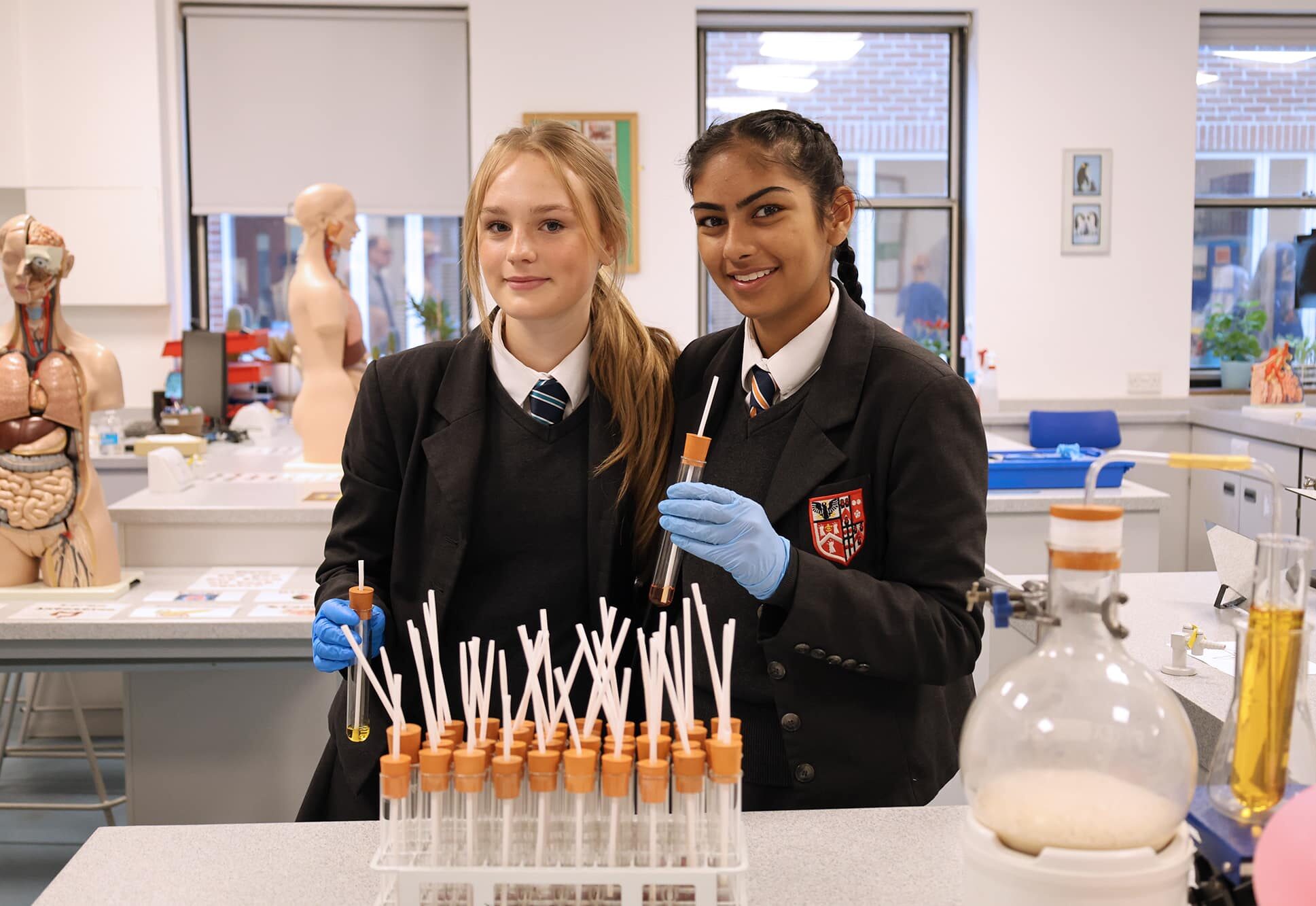 Two students in school uniforms and blue gloves stand in a science lab, holding test tubes and smiling. Join us at our Open Events to explore our labs filled with test tubes, anatomical models, and hands-on experiments.