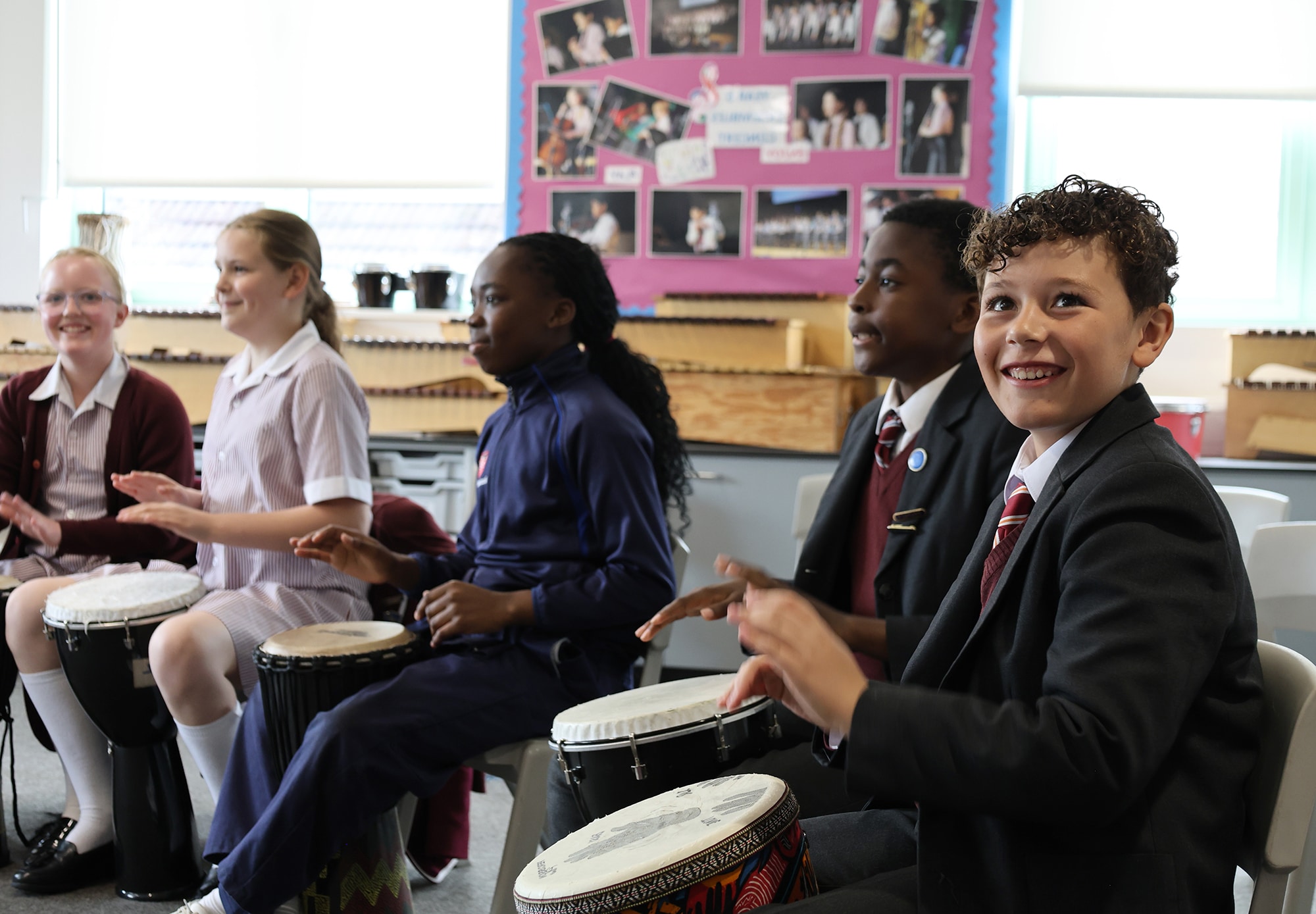 Five schoolchildren sit in a classroom playing hand drums together, smiling and engaged. A colourful noticeboard with photos and text is visible in the background, highlighting upcoming Open Events for families.