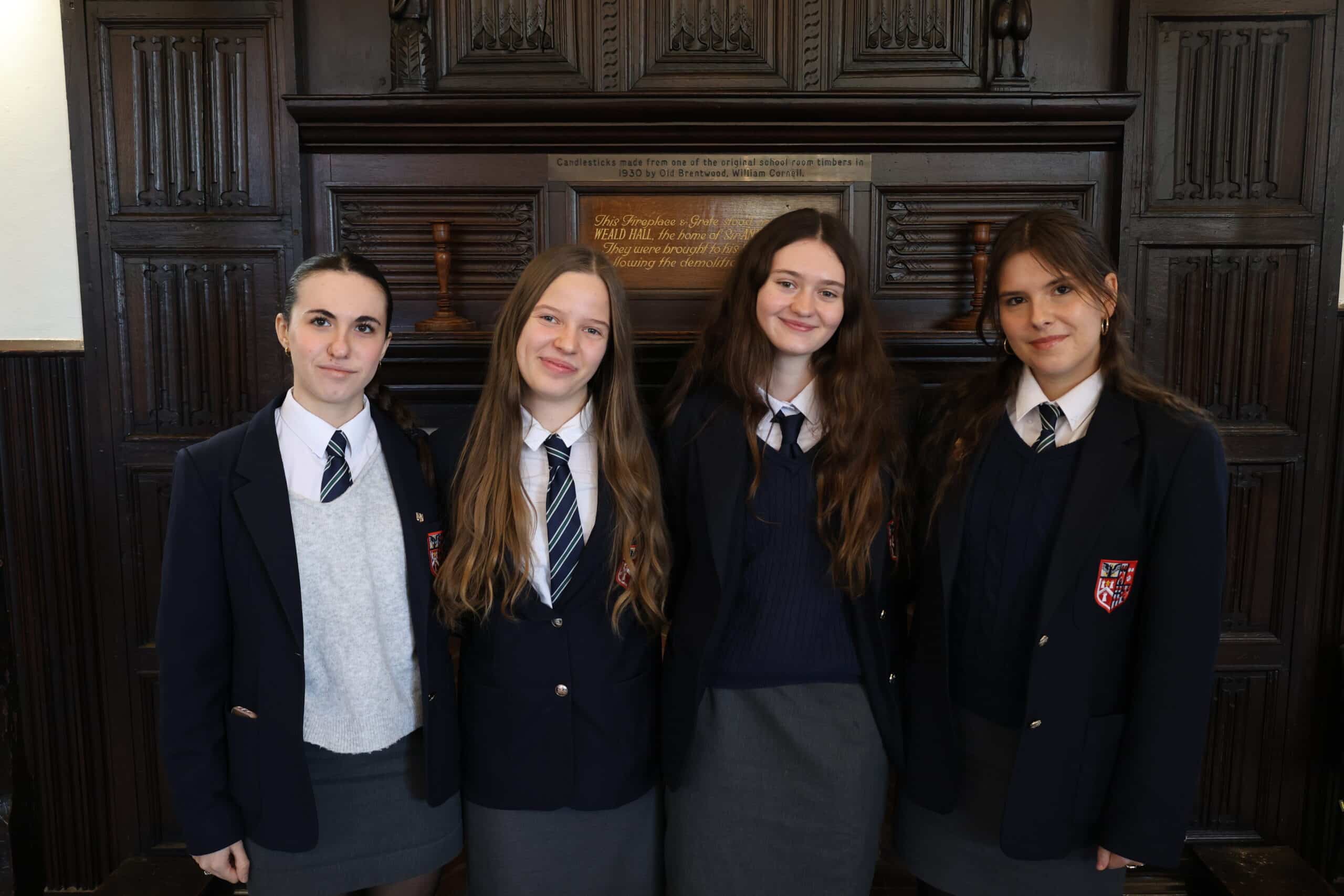 Four students in school uniforms stand indoors, smiling and facing the camera. Dressed in blazers, ties, and skirts, they represent the diversity of academic paths as they stand before dark wood panelling with a plaque above their heads.