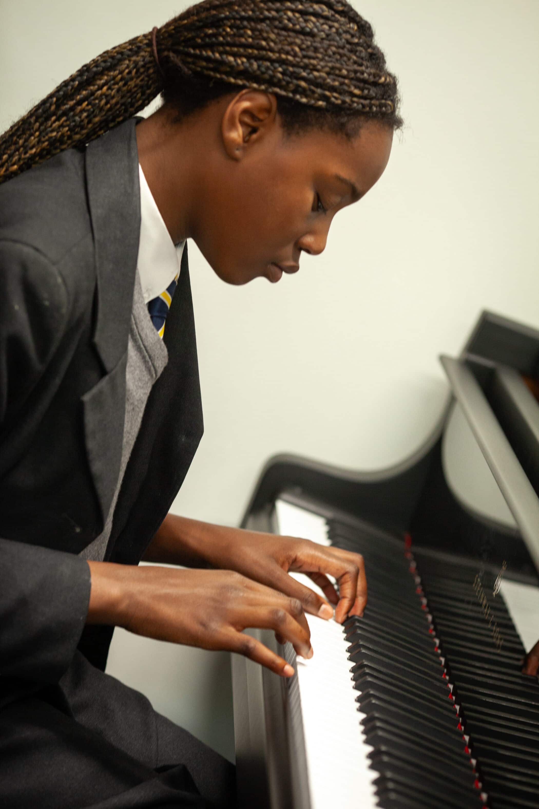 A person wearing a dark suit and tie is playing a grand piano, focused intently on the keys, with plaited hair falling over their shoulder, embodying why music education is important for child development through their dedication and artistry.