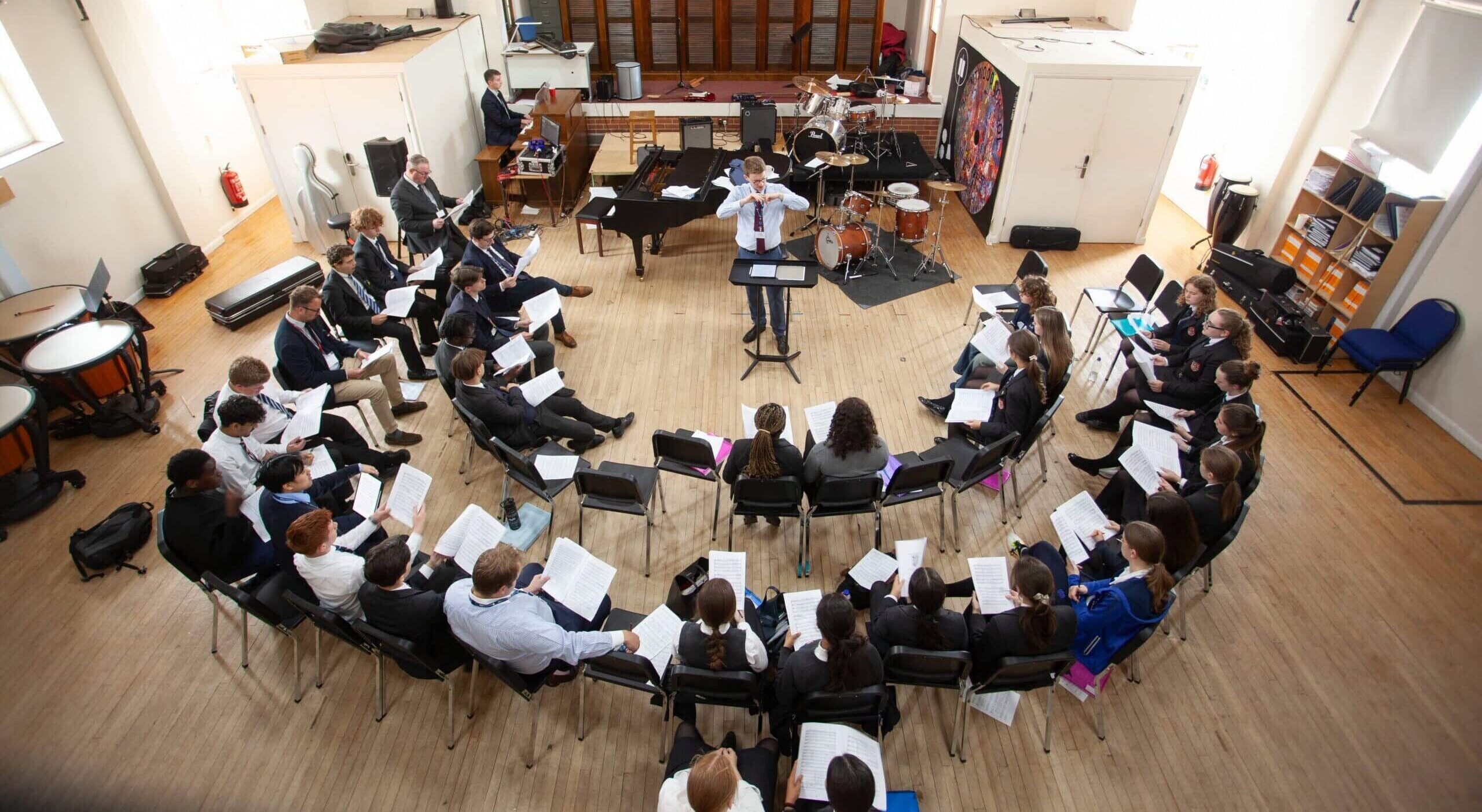 A choir rehearsal in a bright room, with people seated in a semicircle facing a conductor. Musical instruments and sheets are visible, and sunlight streams through the windows.