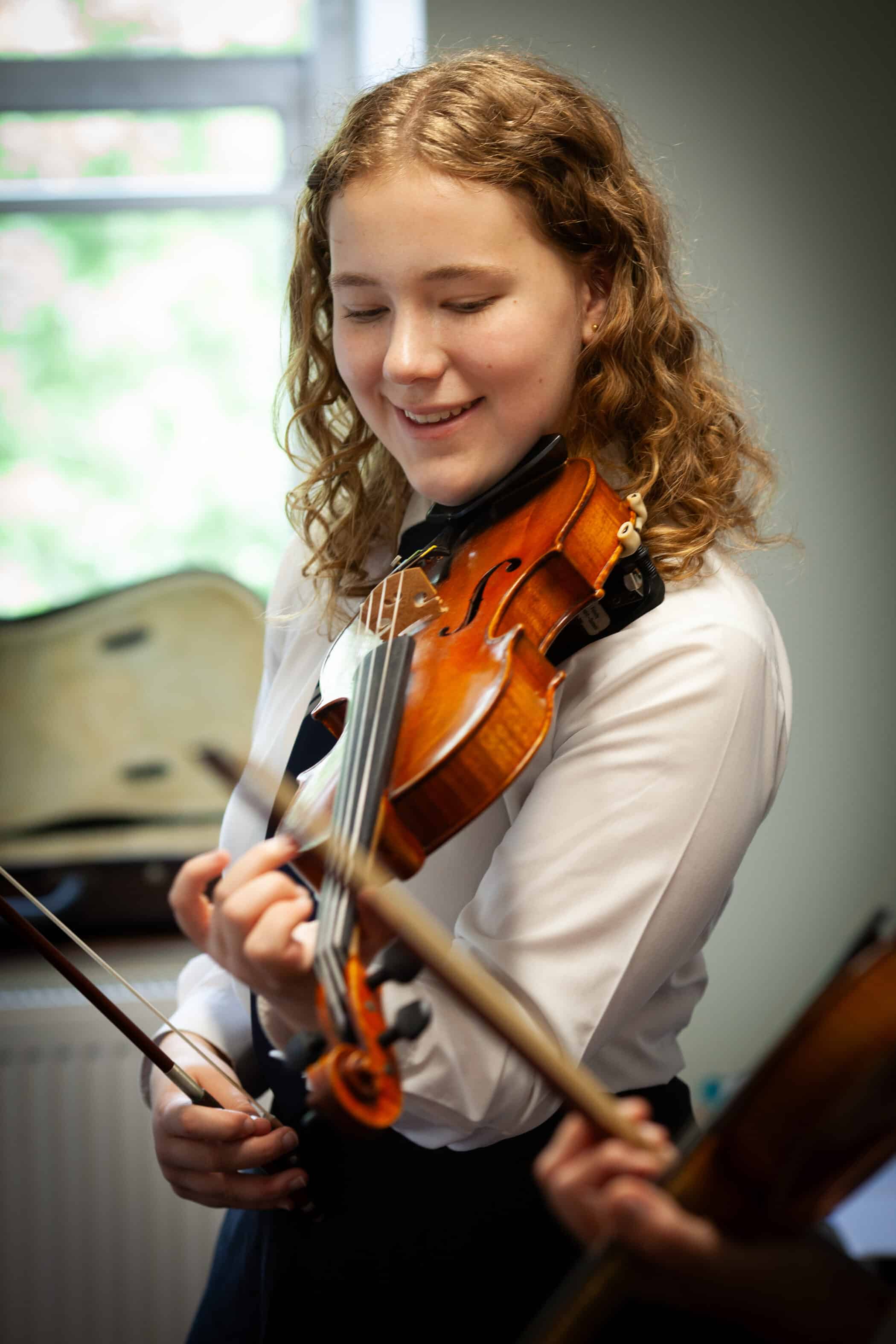 A young person with curly hair smiles while playing a violin indoors, illustrating why music education is important for child development; another violinist is partially visible in the foreground.