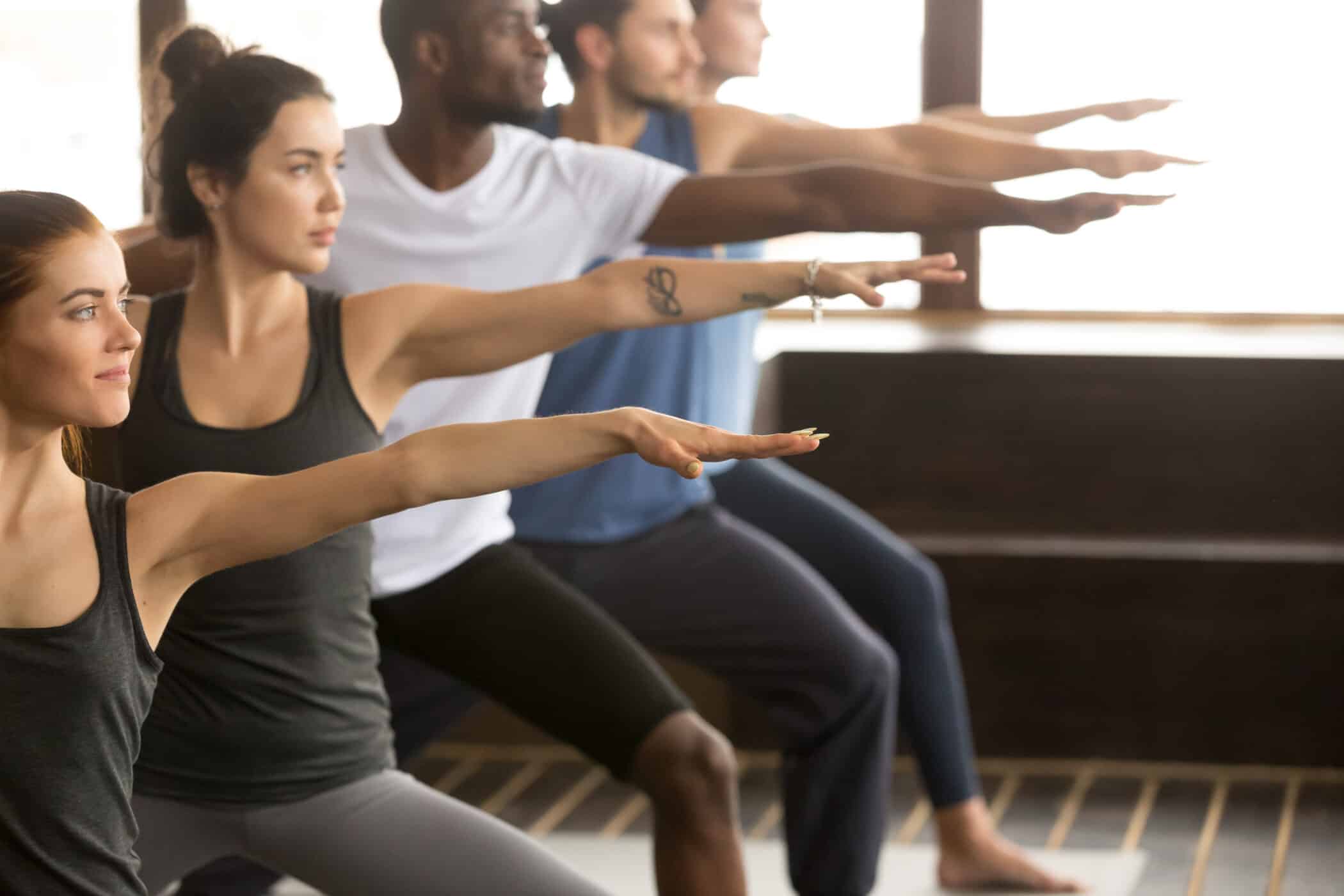 A diverse group of people in sportswear practise the Warrior II yoga pose in a bright studio, standing in a row with arms extended to activate balance and concentration.