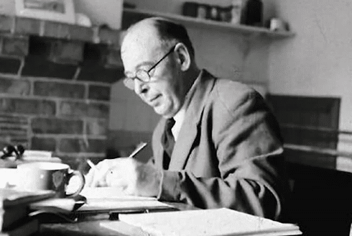 A man wearing glasses and a suit sits at a desk, writing with a pen on paper. Books, cups, and papers are scattered on the desk. A brick fireplace and shelves are visible in the background.